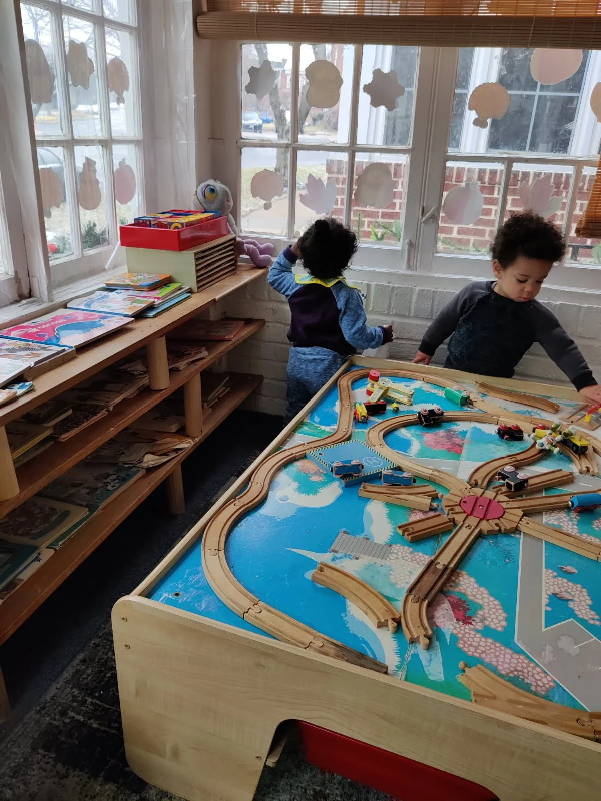 Children playing with a wooden train set at a table in a classroom decorated with paper cloud and star cutouts in the window.