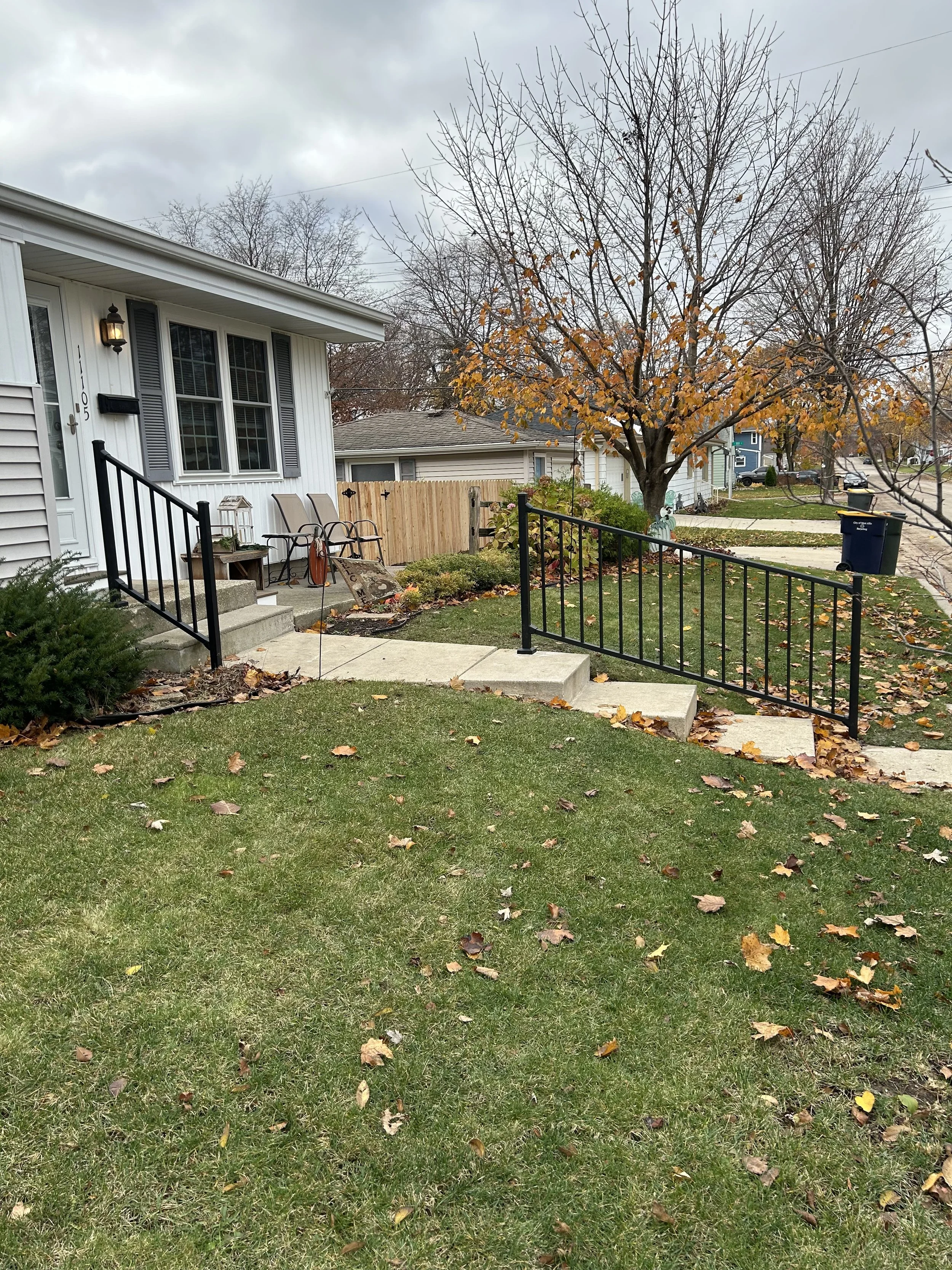 A suburban house with a small front yard, a tree with yellow and orange leaves, fallen leaves on the grass, a concrete sidewalk, black metal railings, and a wooden fence in the background under overcast skies. Cedar fence