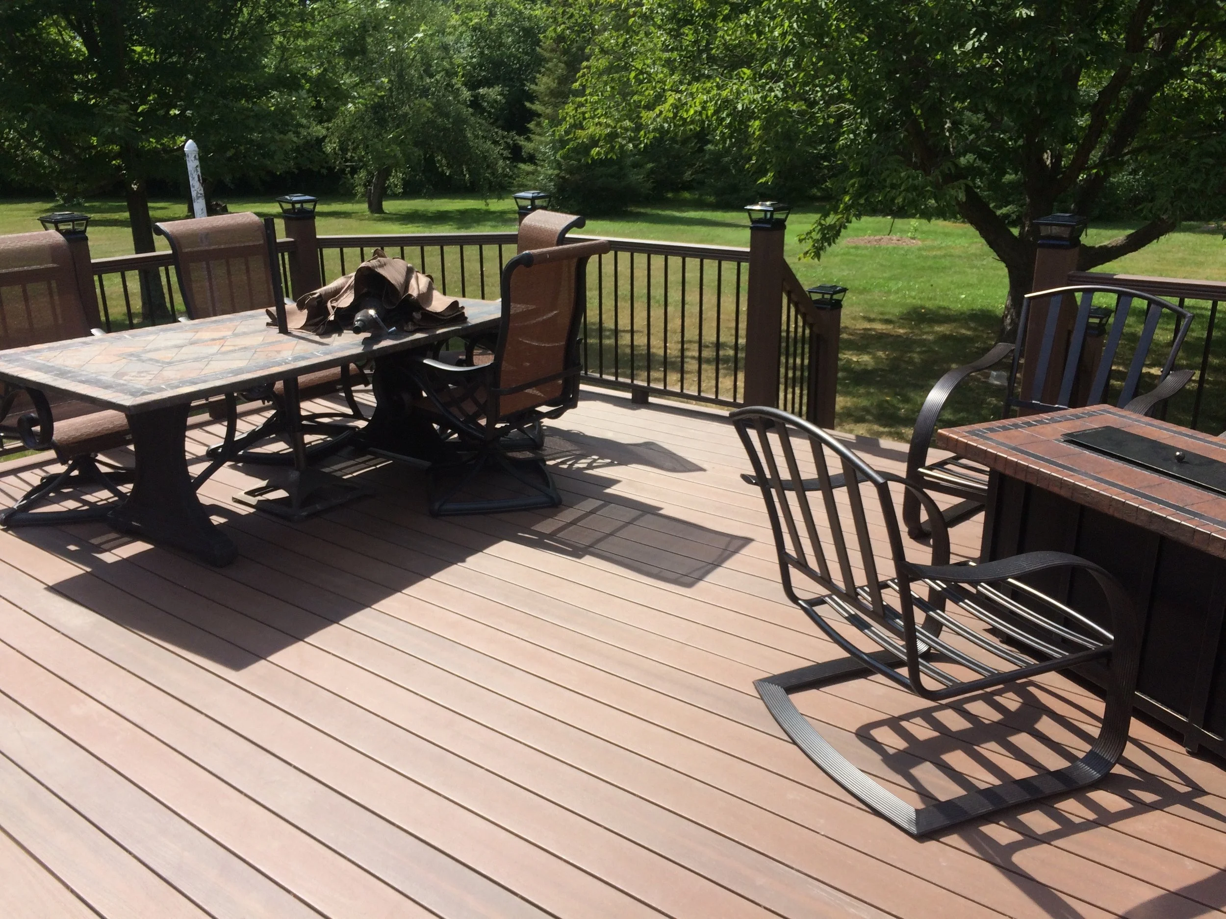 Wooden outdoor composite deck with metal and cushioned chairs, a large dining table with a bag on it, and trees and grass in the background.