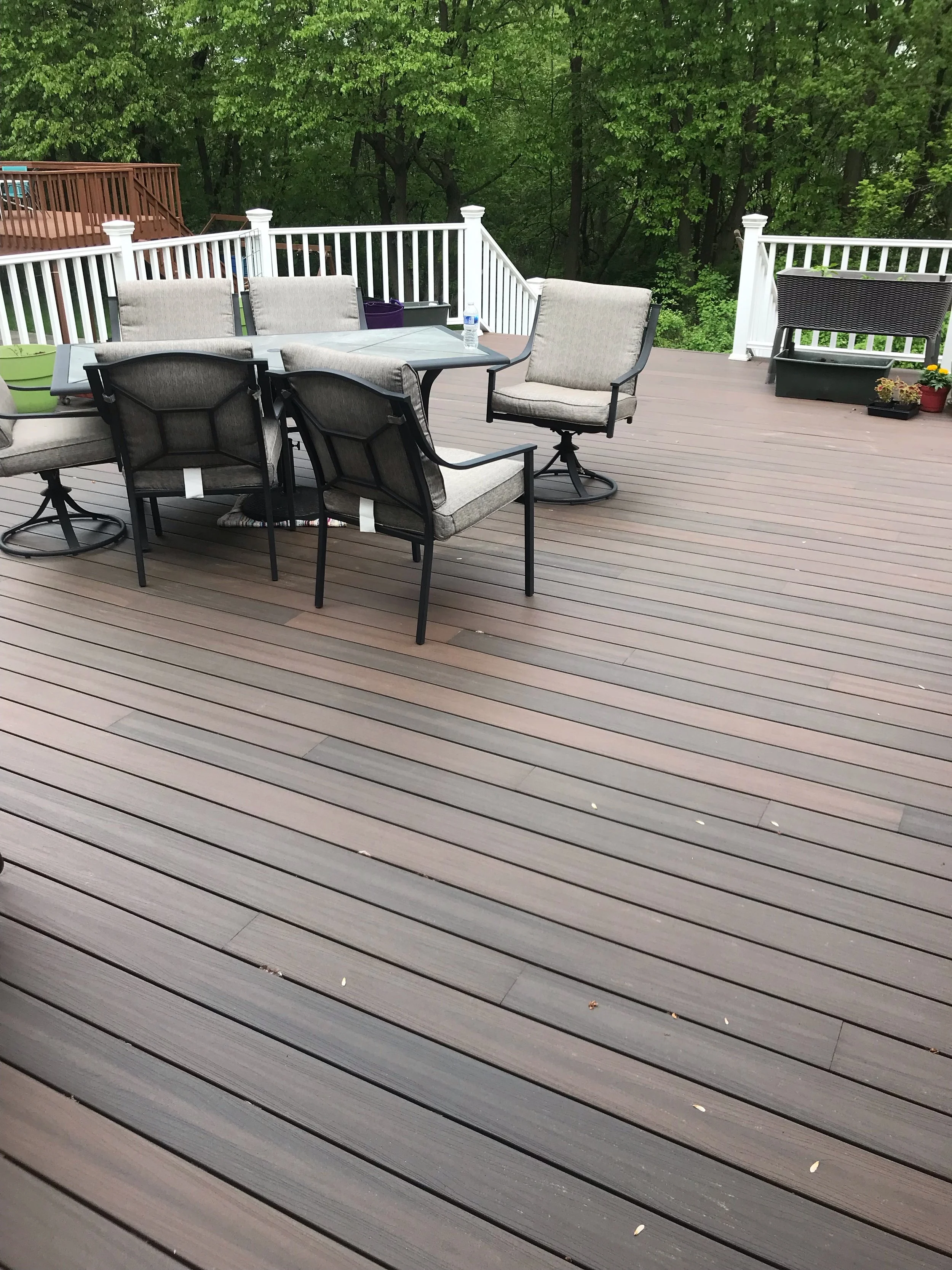Photo of a composite outdoor deck with patio furniture including a table and several chairs, surrounded by green trees.