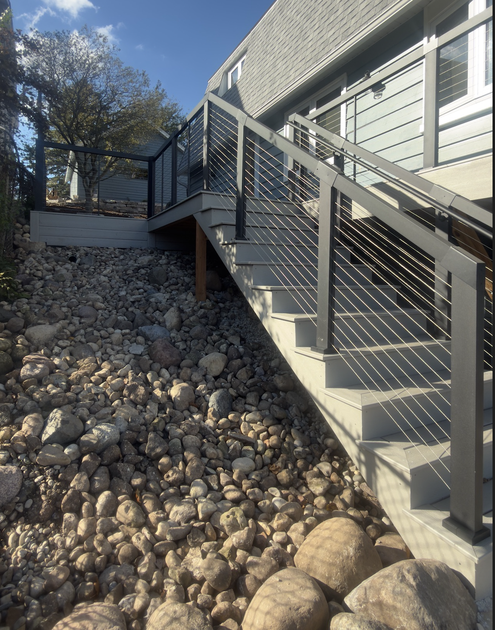 Exterior view of a house with a staircase leading up to a balcony, surrounded by rocks and trees. Composite decking with aluminum cable railing.