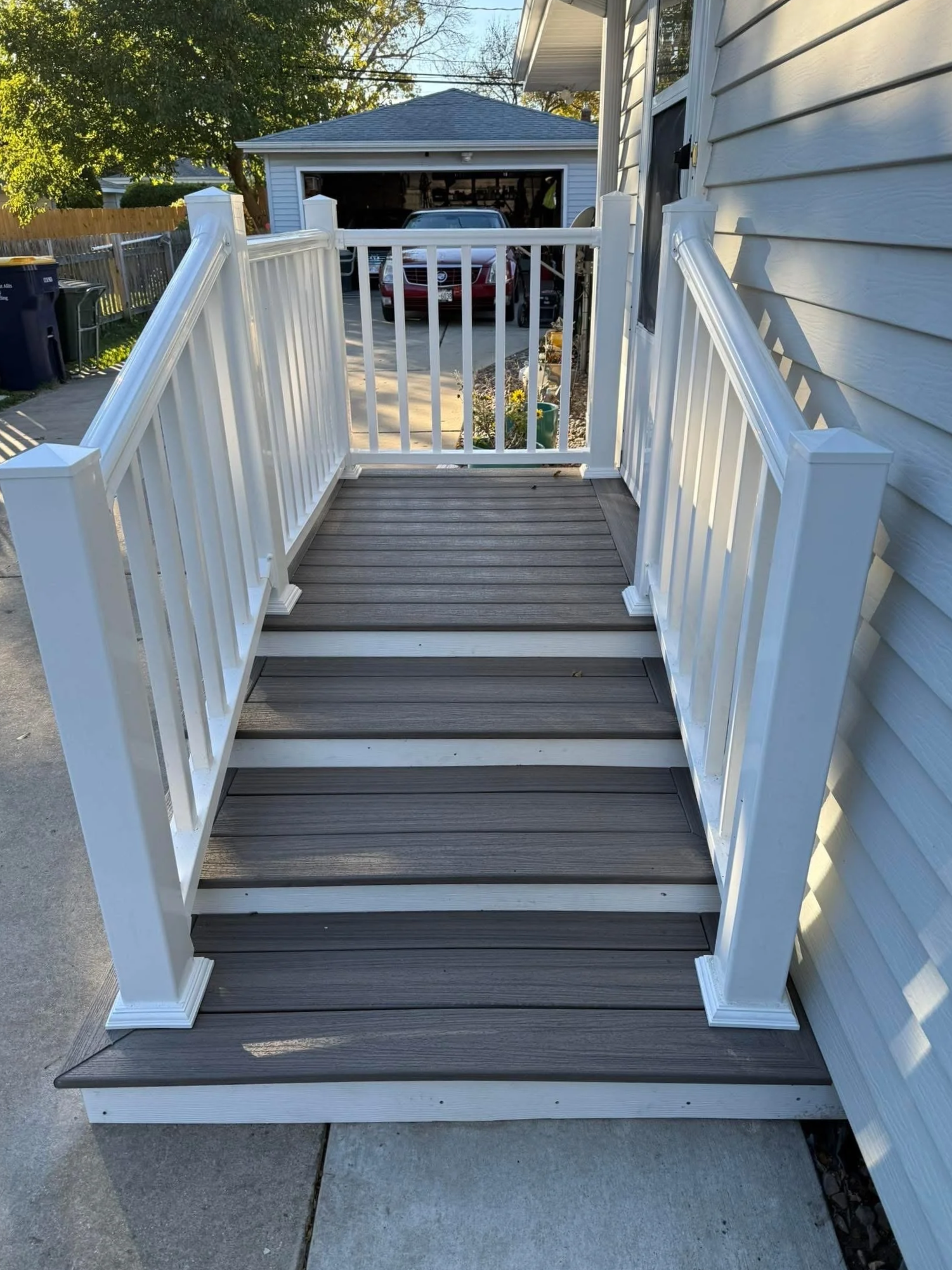 A newly built small porch with white railings and brown composite decking, attached to a house with beige siding. A backyard with a driveway, garage, parked red car, and trees is visible in the background.