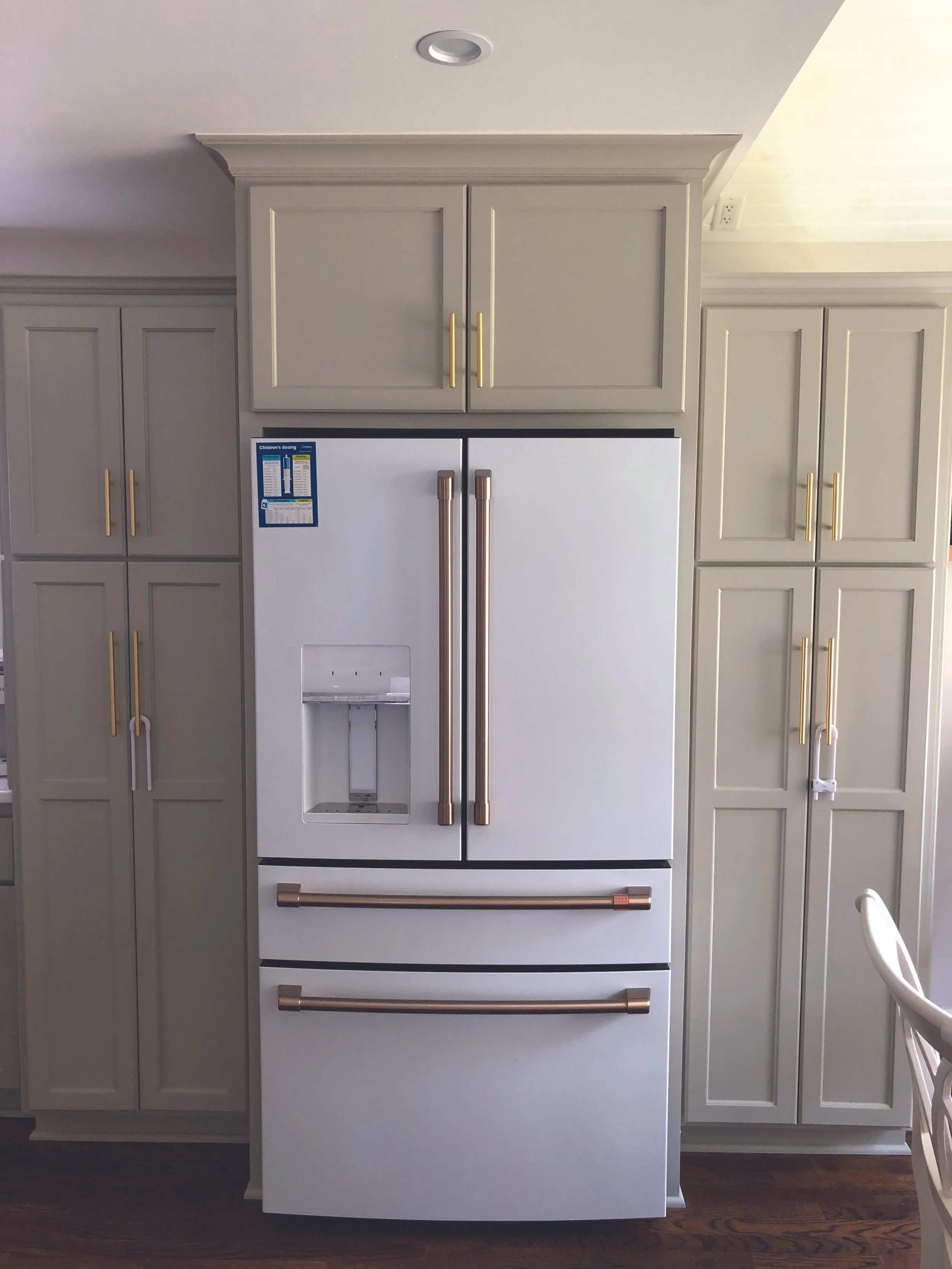 A white refrigerator with gold handles in a kitchen, surrounded by light gray cabinets with gold handles, and a wooden floor.
