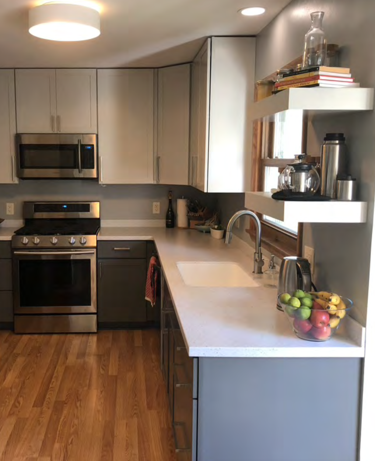 Modern kitchen with gray cabinets, stainless steel appliances, a white countertop, hardwood floor, open shelving with books and kitchen items, and a bowl of mixed apples and bananas.