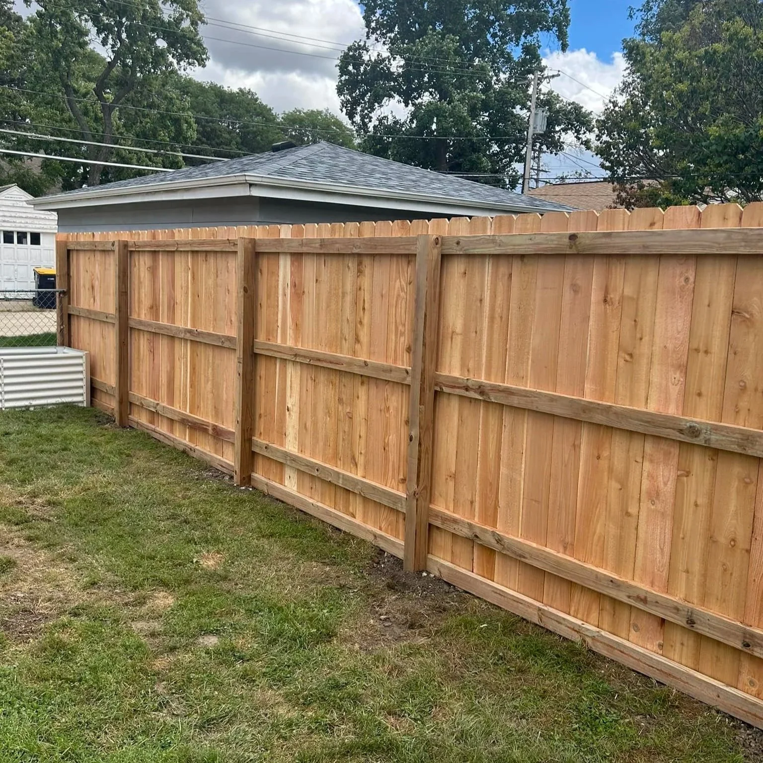 A new wooden cedar dog ear fence installed in a backyard, with a grass lawn and a small white garden bed in the corner. A house with a gray roof and some trees are visible in the background.