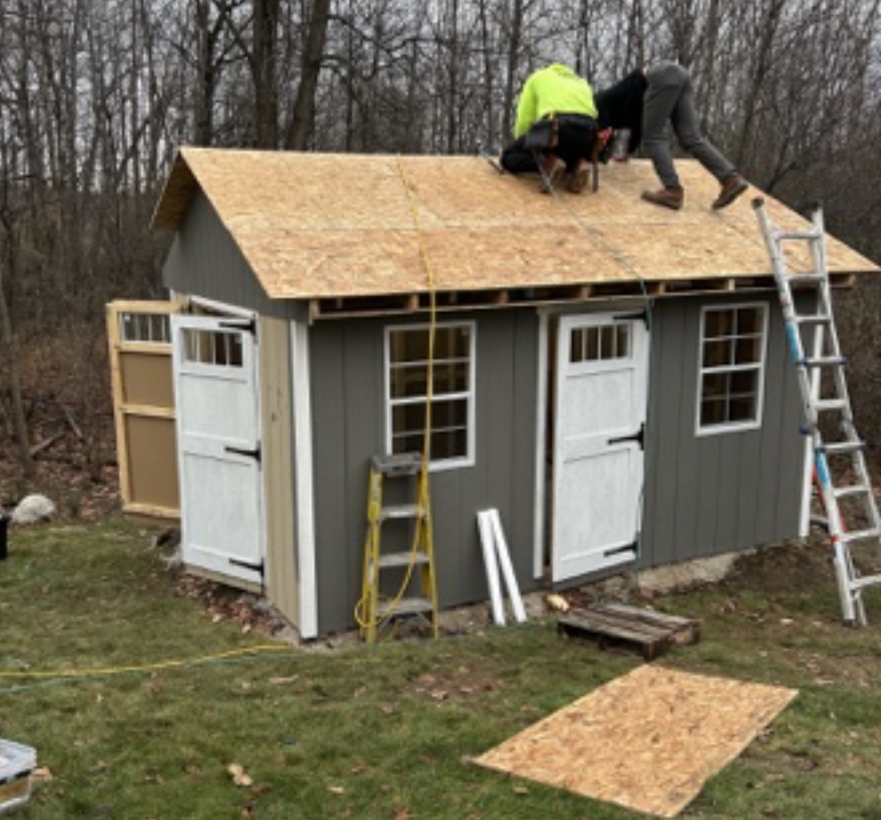 Two people working on installing a shingled roof on a small gray shed with two white doors and two windows, surrounded by ladders and construction tools in a yard with trees in the background.