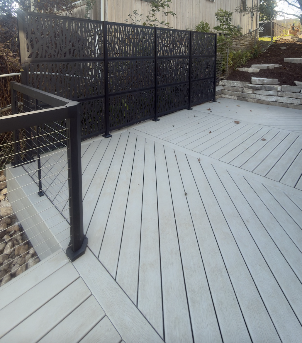 Newly installed black decorative privacy fence on a light-colored composite deck with a small garden area and stone steps in the background.
