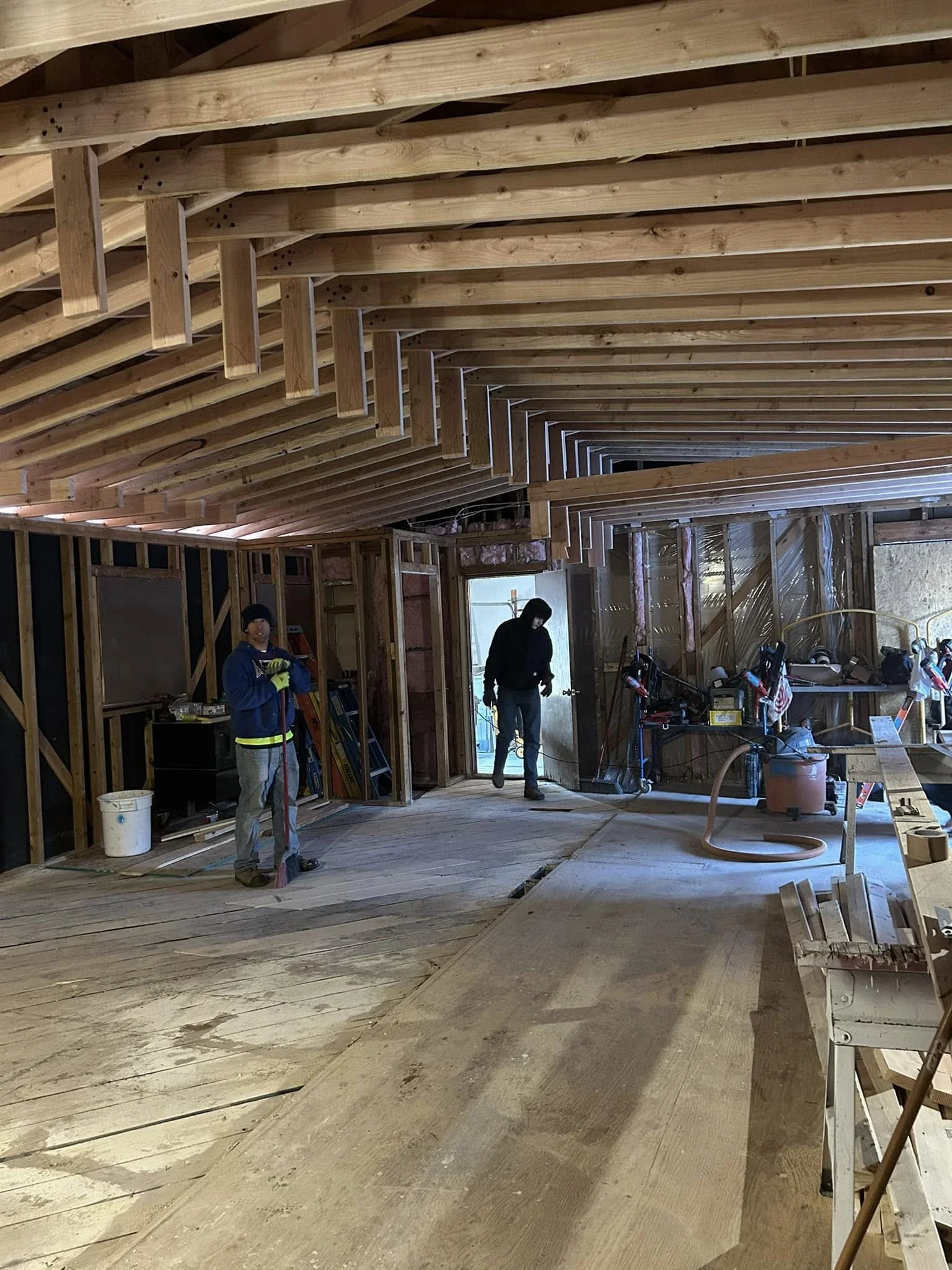 Construction workers inside a house under construction, with exposed wooden framing, a sloped ceiling, and tools scattered around.