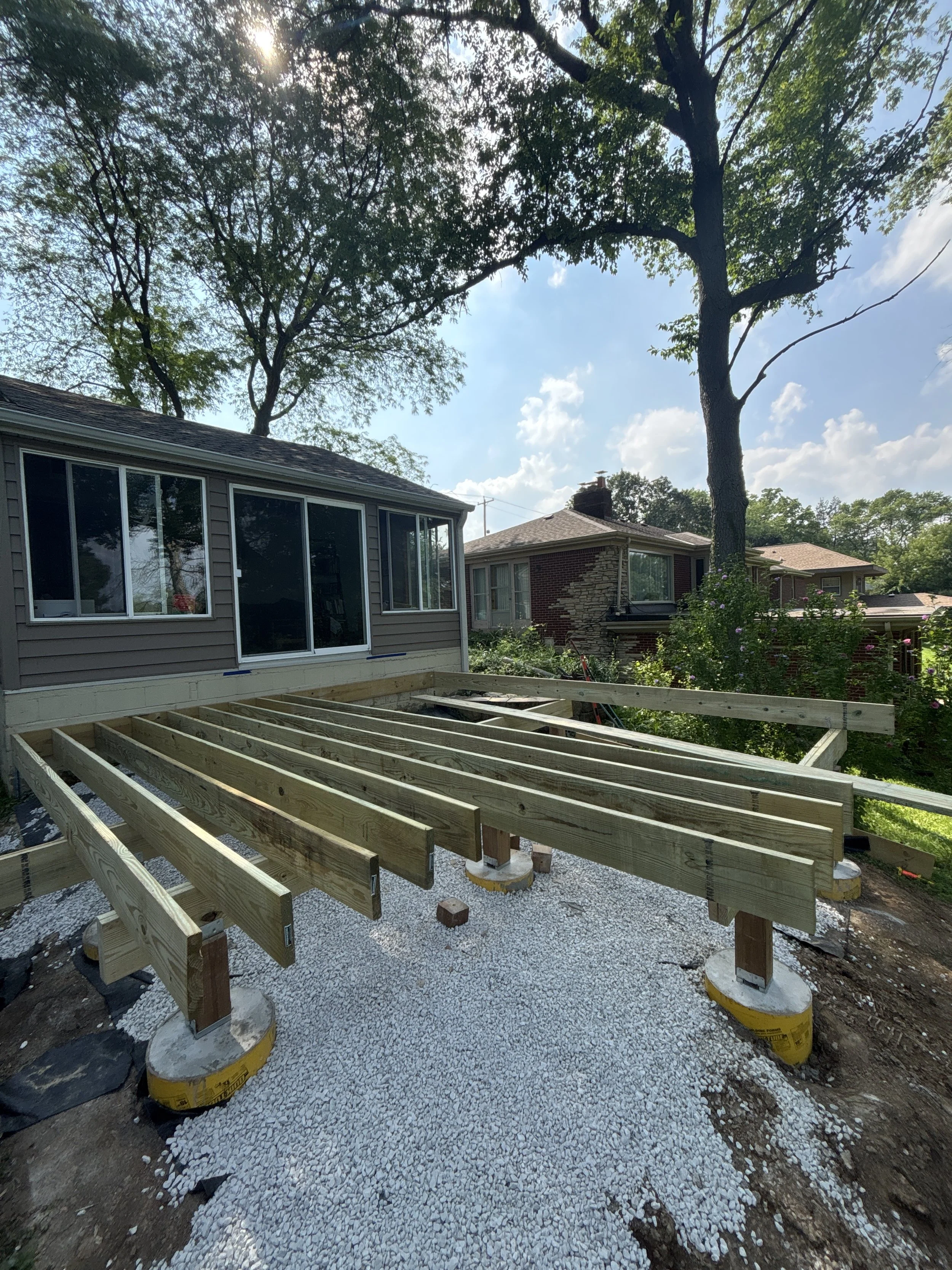 Construction of a deck in progress on the backyard of a house, with wooden beams and supports on a gravel base, surrounded by trees and neighboring houses under a partly cloudy sky.