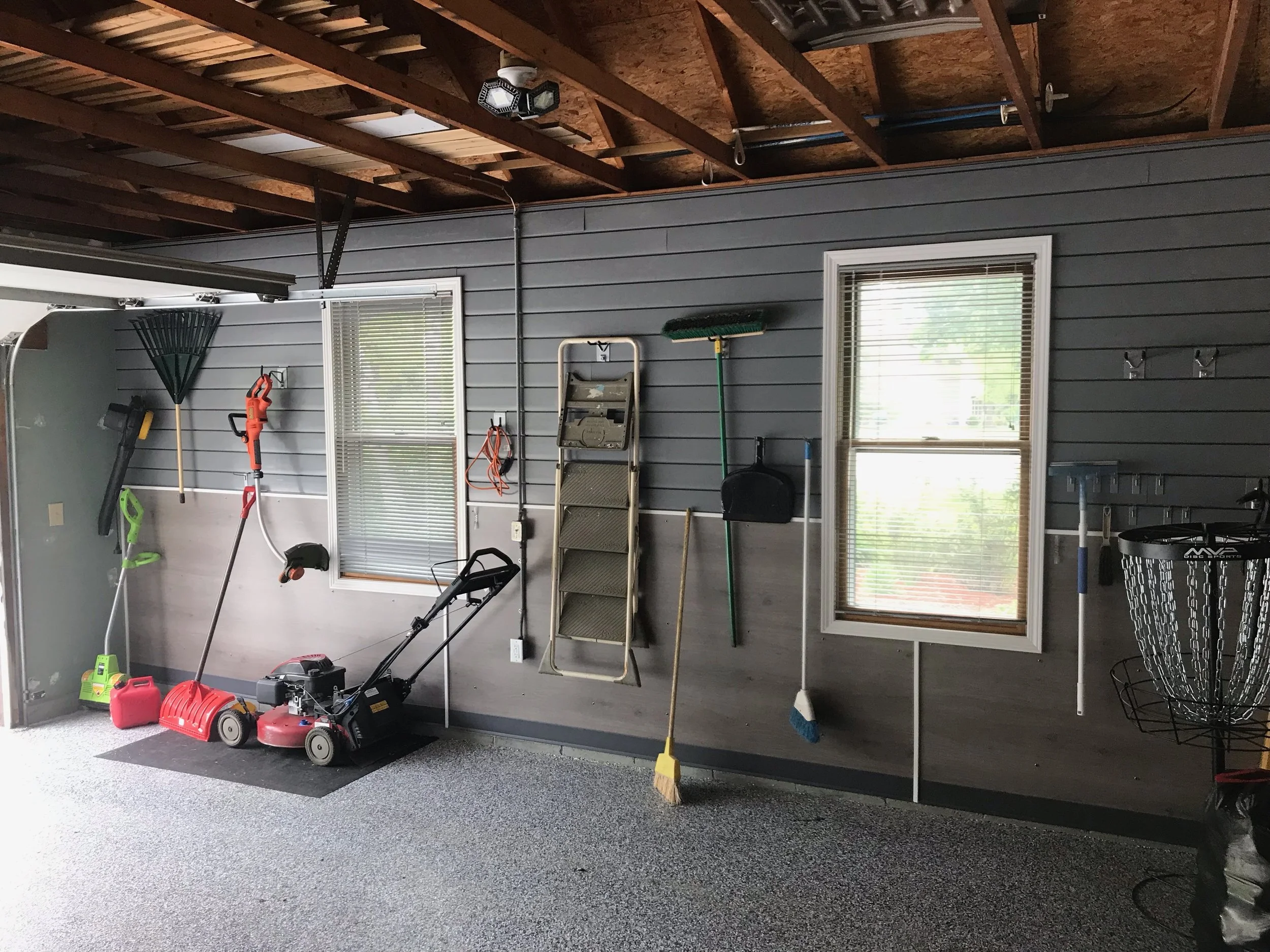 A garage wall with various gardening tools, including a rake, a leaf blower, a lawnmower, and a snowblower, hanging by the windows. There are two windows with blinds, a foldable step ladder, and a window with hooks on the wall. The garage has a textu
