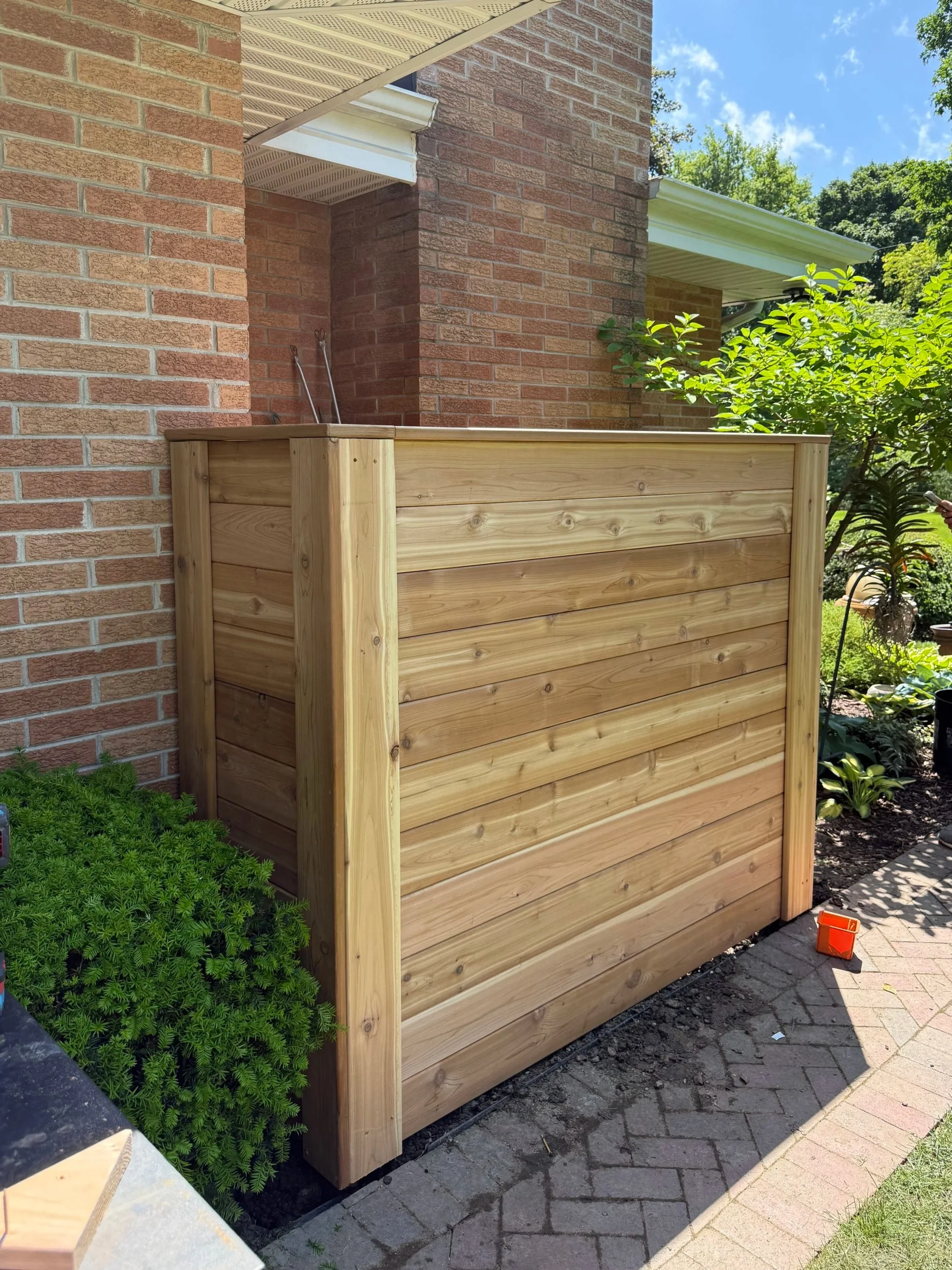 A newly built wooden cedar horizontal board privacy fence panel placed near a brick house, with plants and a brick pathway nearby, under a blue sky.
