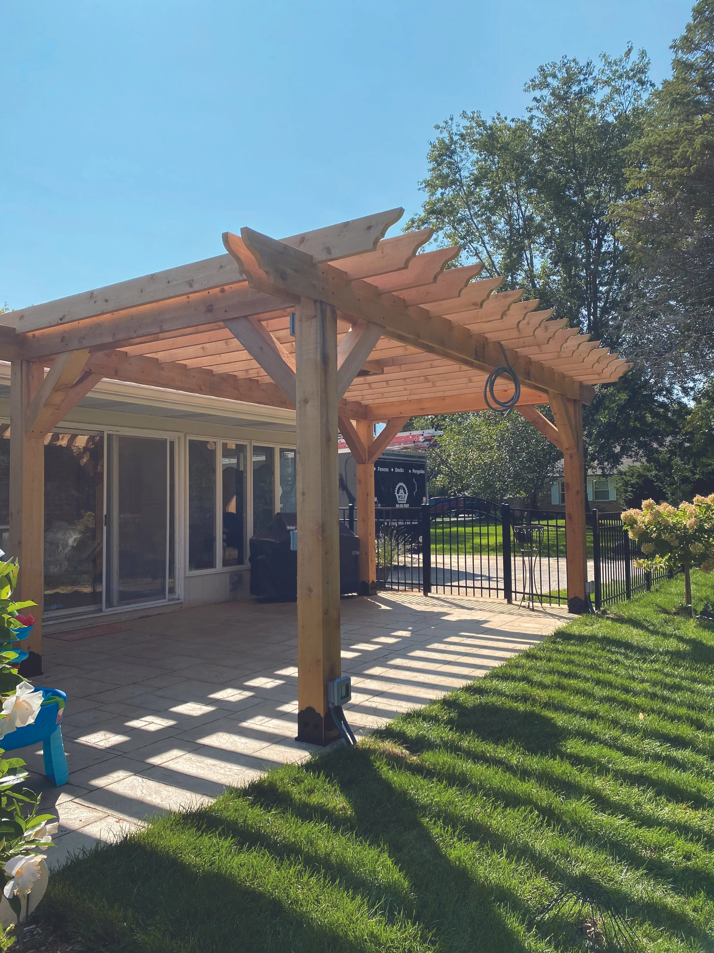 Backyard with wooden pergola over patio, black metal gate, sliding glass doors, green grass, and trees in background.