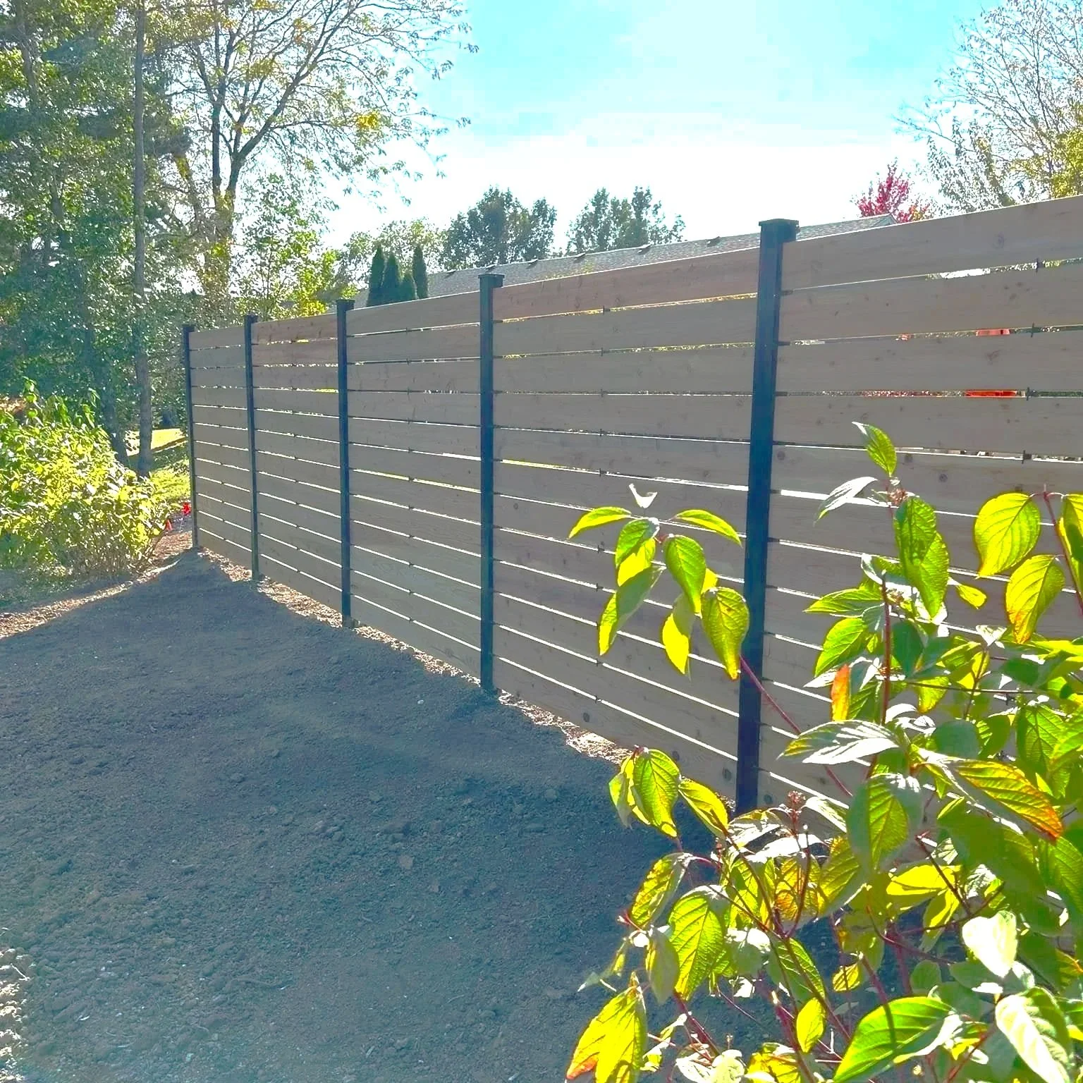A cedar-tone wooden fence with horizontal planks and aluminum posts in a backyard, with green foliage and trees in the background and sunlight shining through.