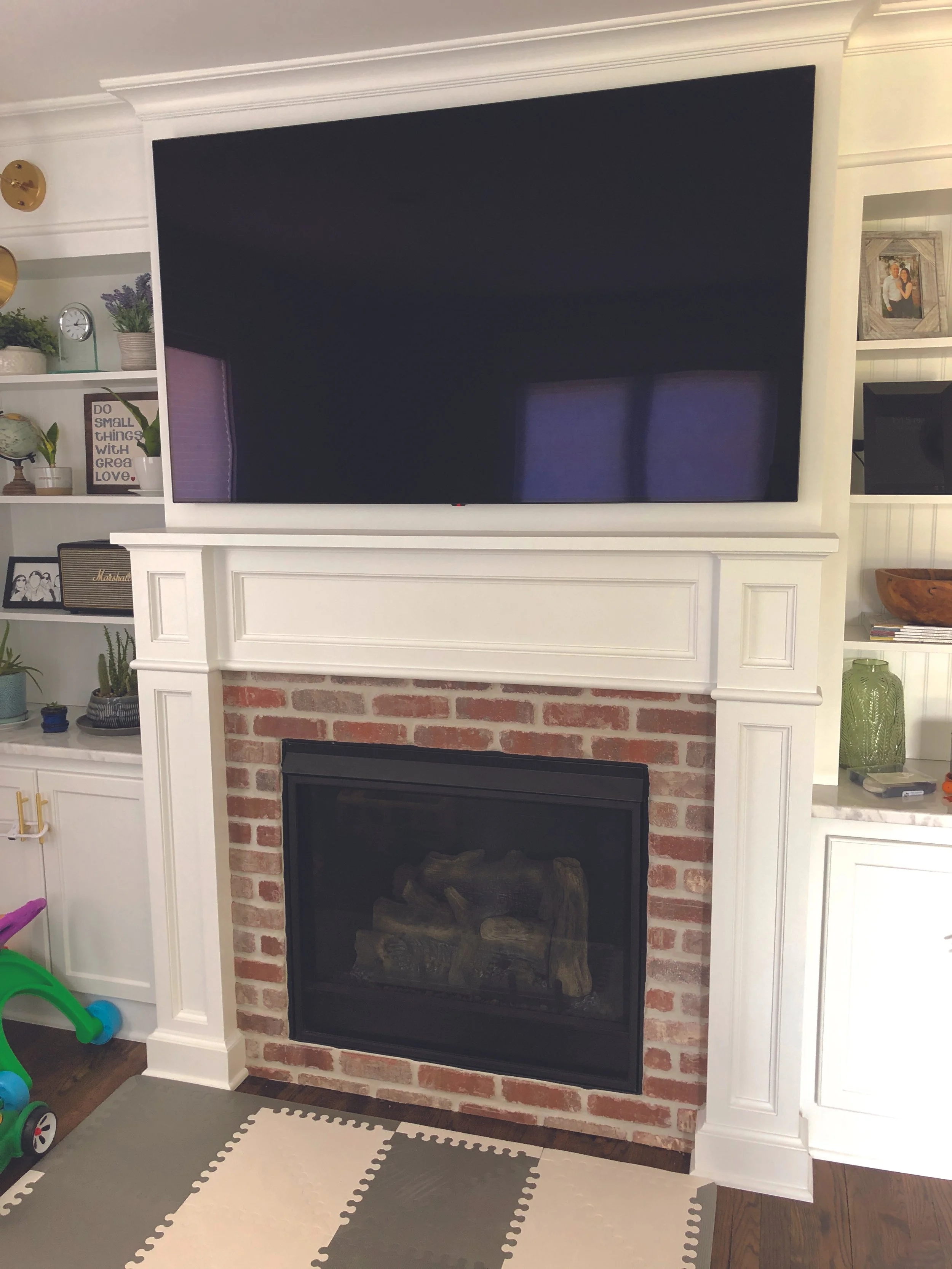A living room fireplace with a brick surround and white wood mantel, topped by a large flat-screen TV. To the left are white shelves with decorative items and framed photos. To the right, a green vase and smaller decor pieces are visible.