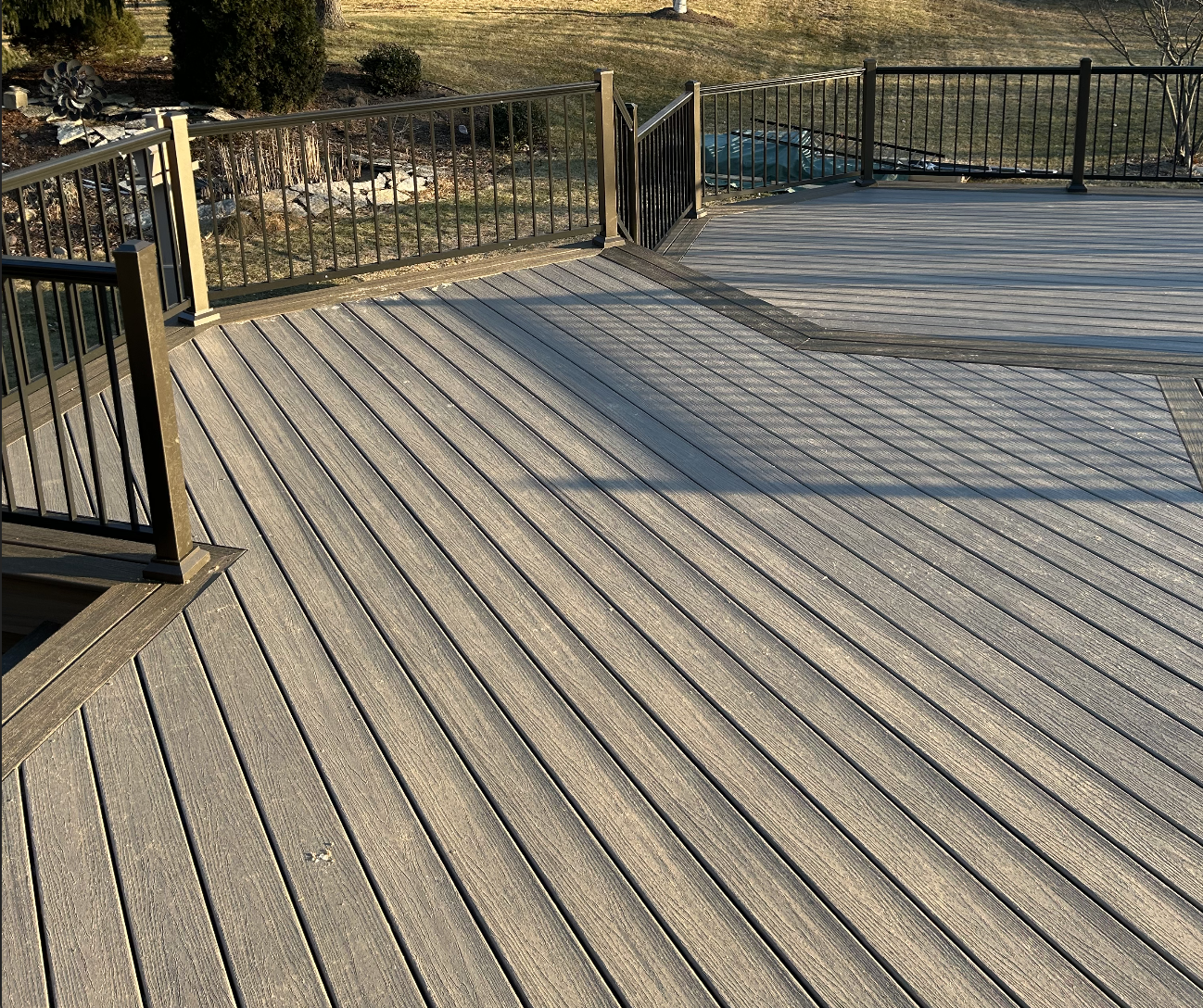 wooden deck with metal railing, overlooking a grassy yard and garden area with trees and shrubs.