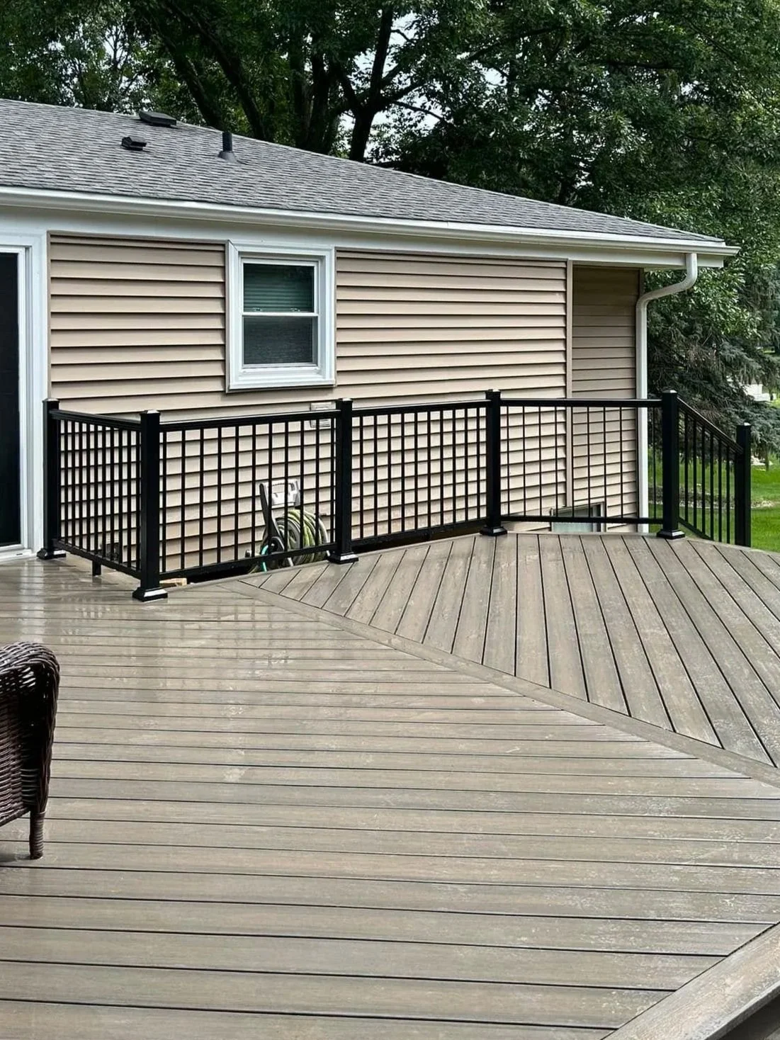 A composite deck with a black aluminum railing attached to a house with beige vinyl siding and a small window. The deck appears wet, and there is a garden hose stored near the house. Trees are visible in the background.