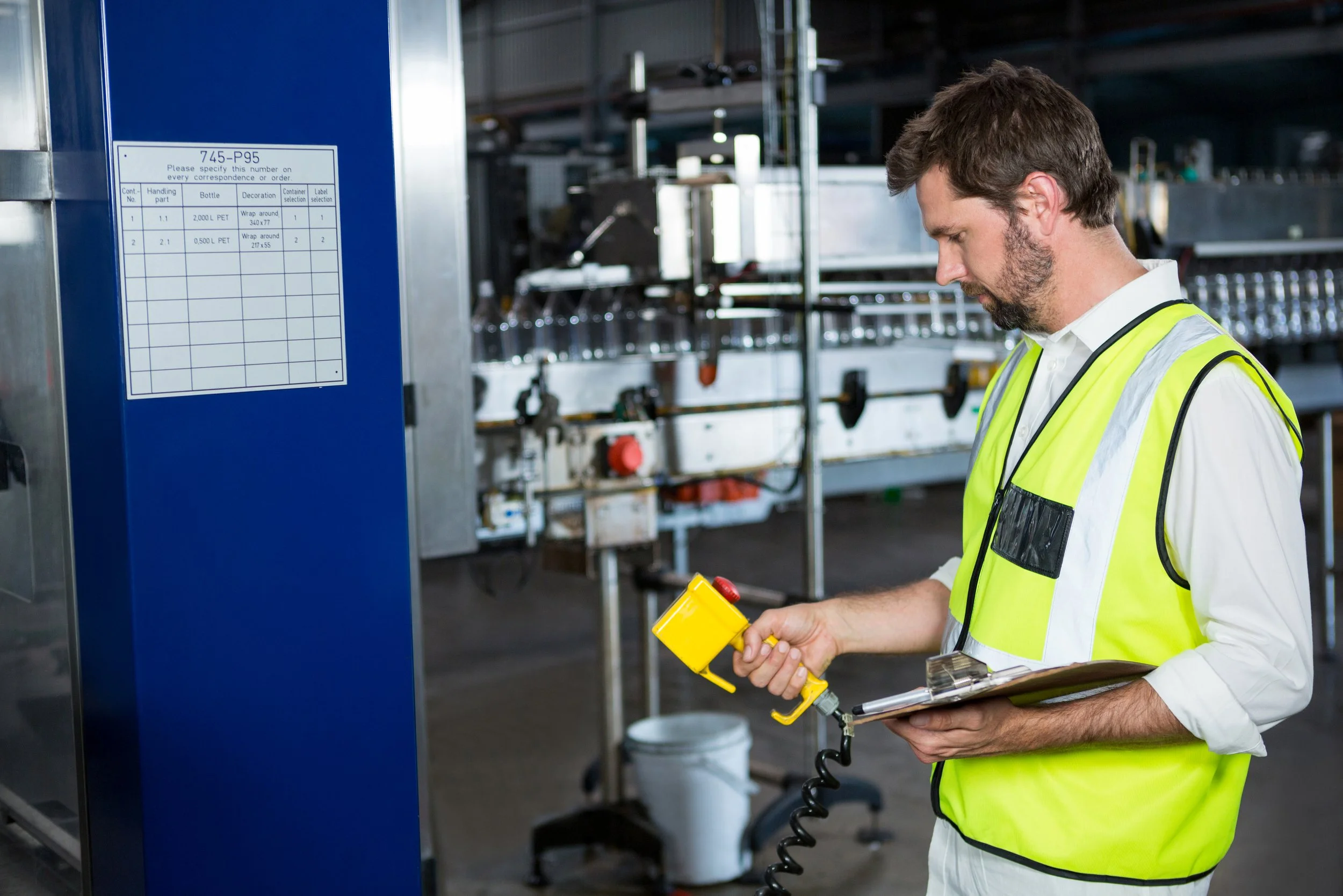 A man wearing a yellow safety vest holds a clipboard and a handheld scanner in an industrial or manufacturing setting.