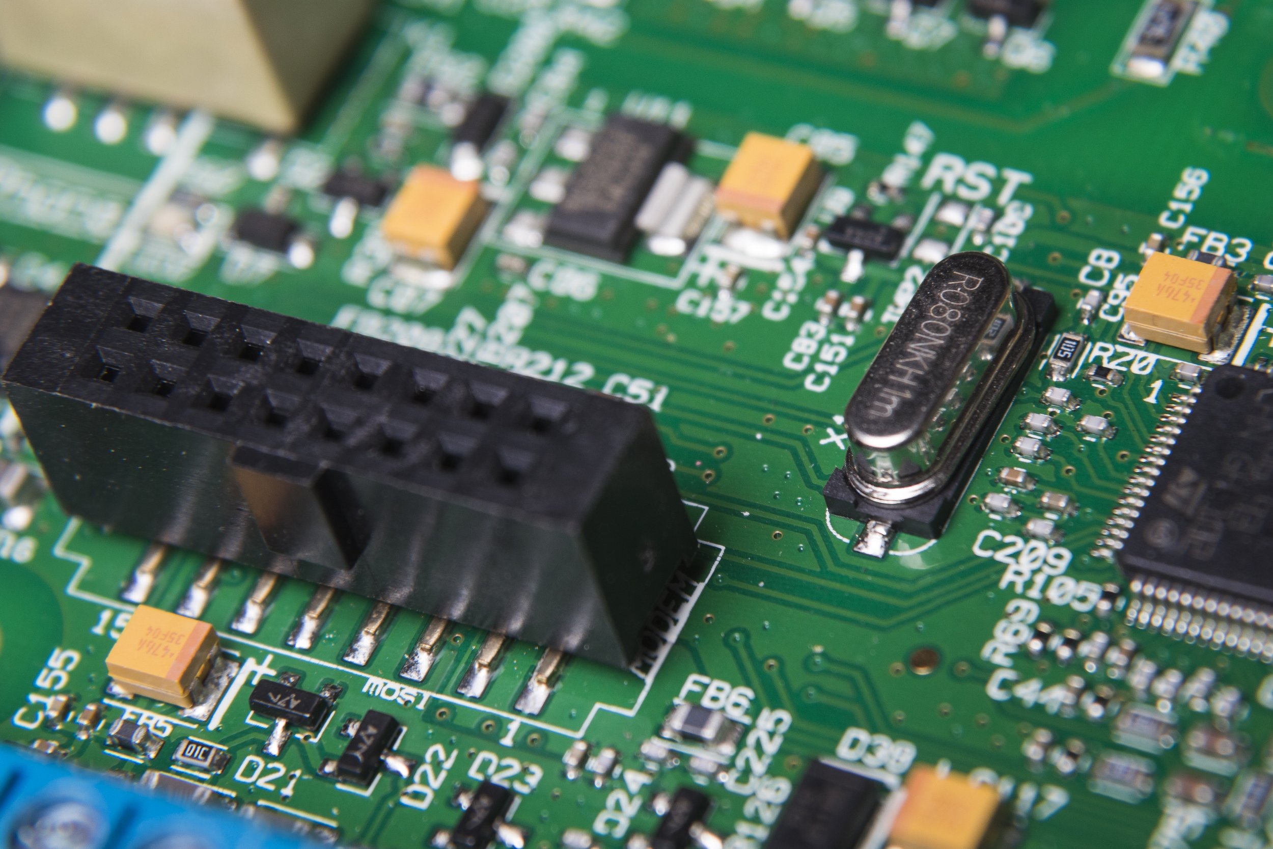 Close-up of a green circuit board with various black, yellow, and orange electronic components, including a black connector, a silver crystal oscillator, and numerous small surface-mount resistors and capacitors.