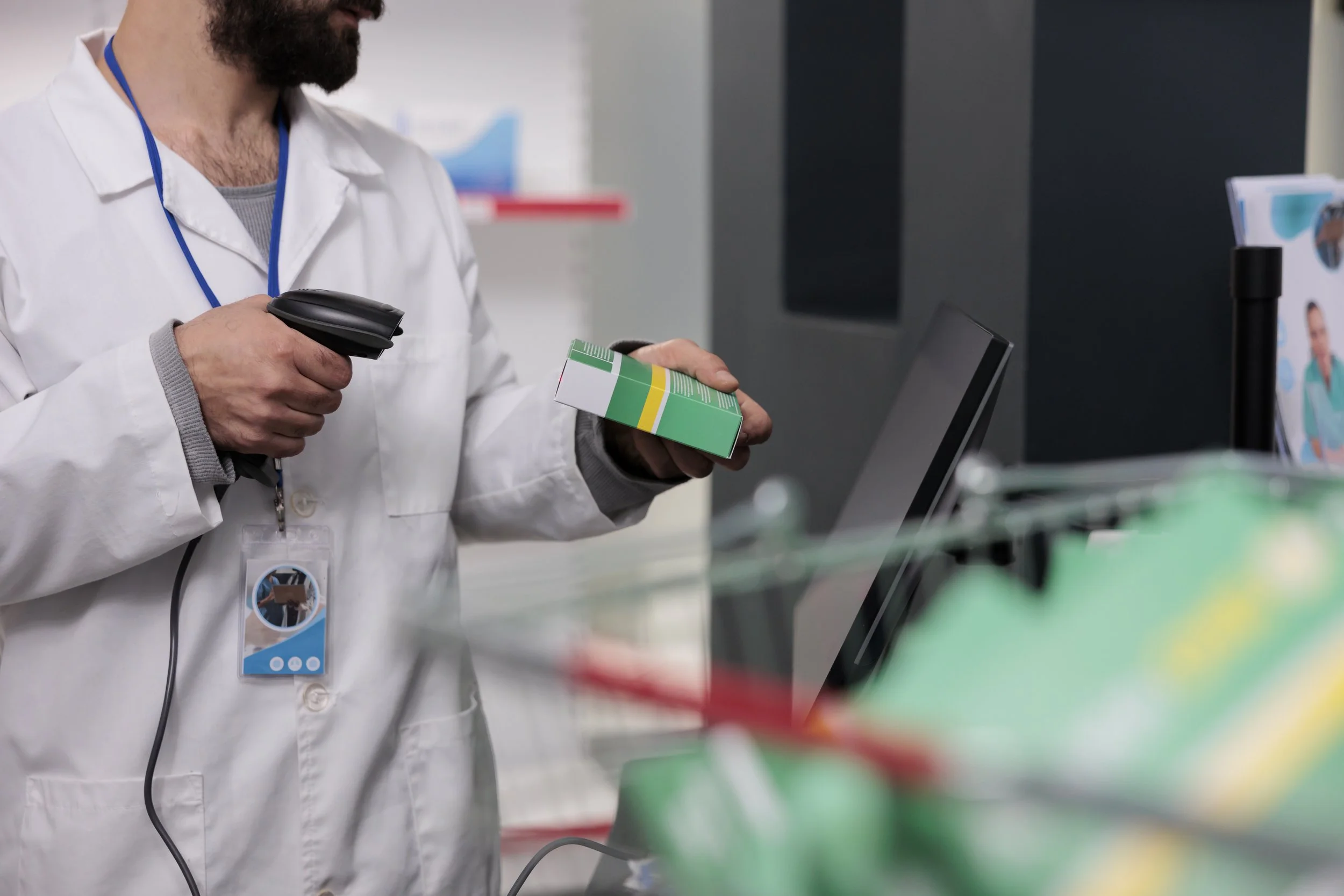 A healthcare worker wearing a white coat and a blue badge scanner is holding a medicine box in one hand and a barcode scanner in the other, in a medical setting with a computer and medical supplies in the background.
