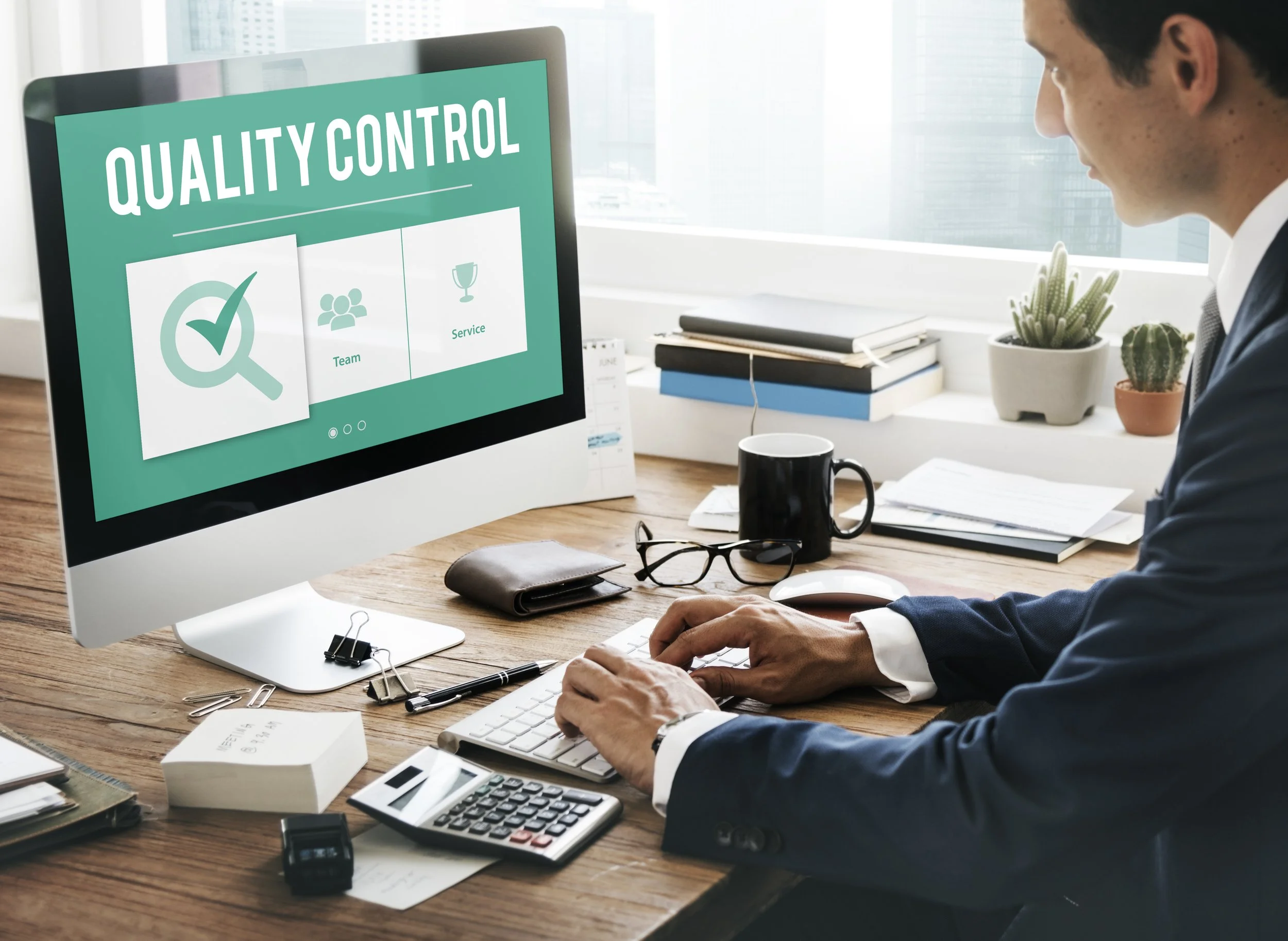 A man in a business suit working at a desk with a computer monitor displaying a 'Quality Control' screen, surrounded by office supplies and potted plants.