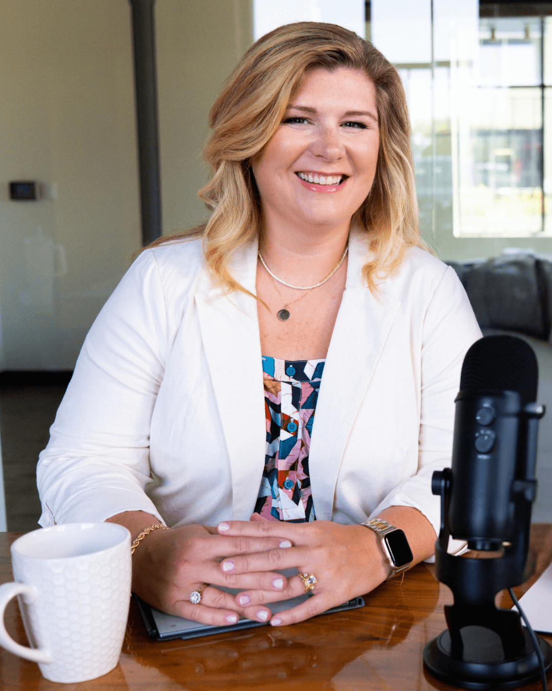 Lauren Cockerell, business owner and PR strategist, smiles while sitting at a wooden desk with a microphone, coffee mug, and notebook, preparing to record a podcast episode.