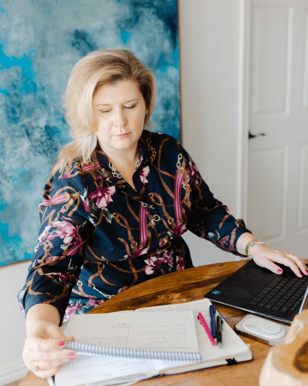 Lauren Cockerell, business strategist and podcast host, reviews marketing materials and works on her laptop at a wooden desk with colorful artwork behind her.