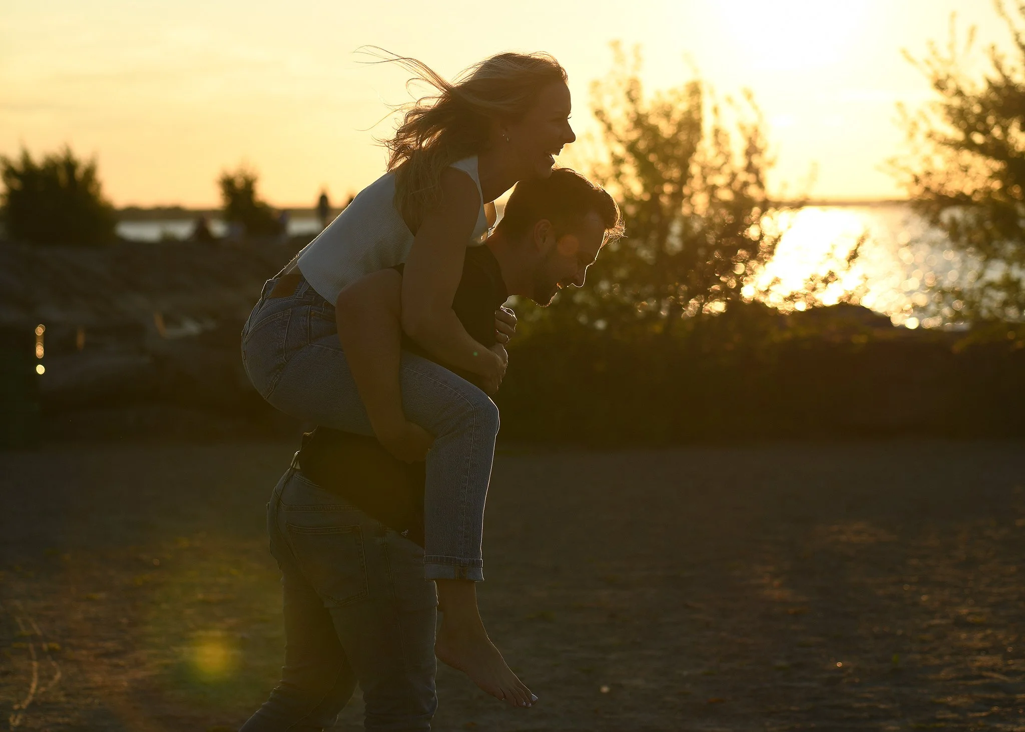 A person carrying another person on their back at sunset, near a body of water, with trees in the background.