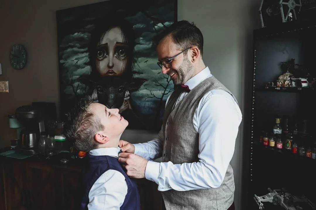 A man in a vest and bow tie helping a young boy adjust his suit in a room with a dark, surreal painting and a shelf with various items.