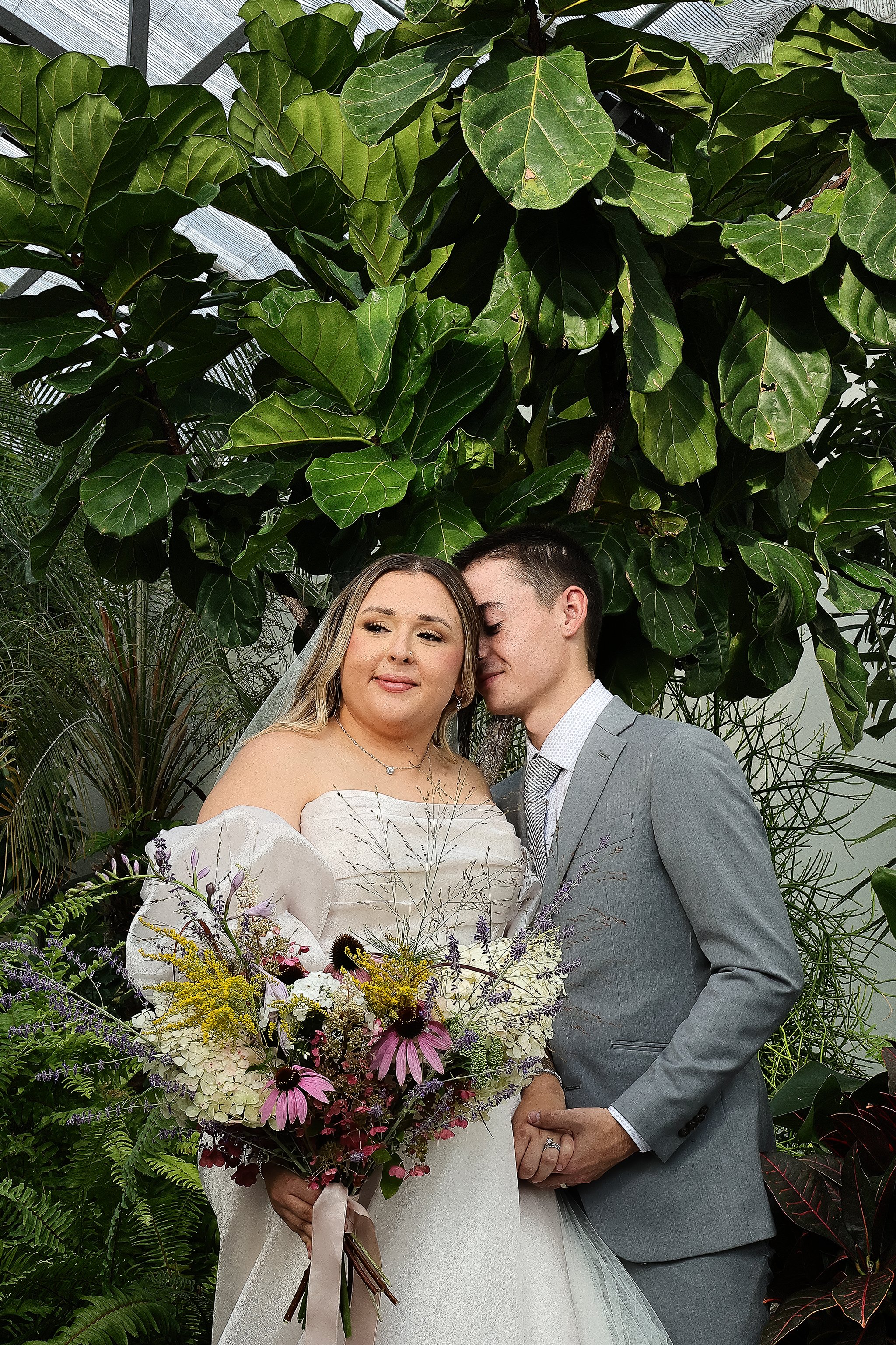 A couple in wedding attire stand under green foliage. The bride holds a colorful bouquet of flowers, and the groom leans towards her, wearing a gray suit and tie.