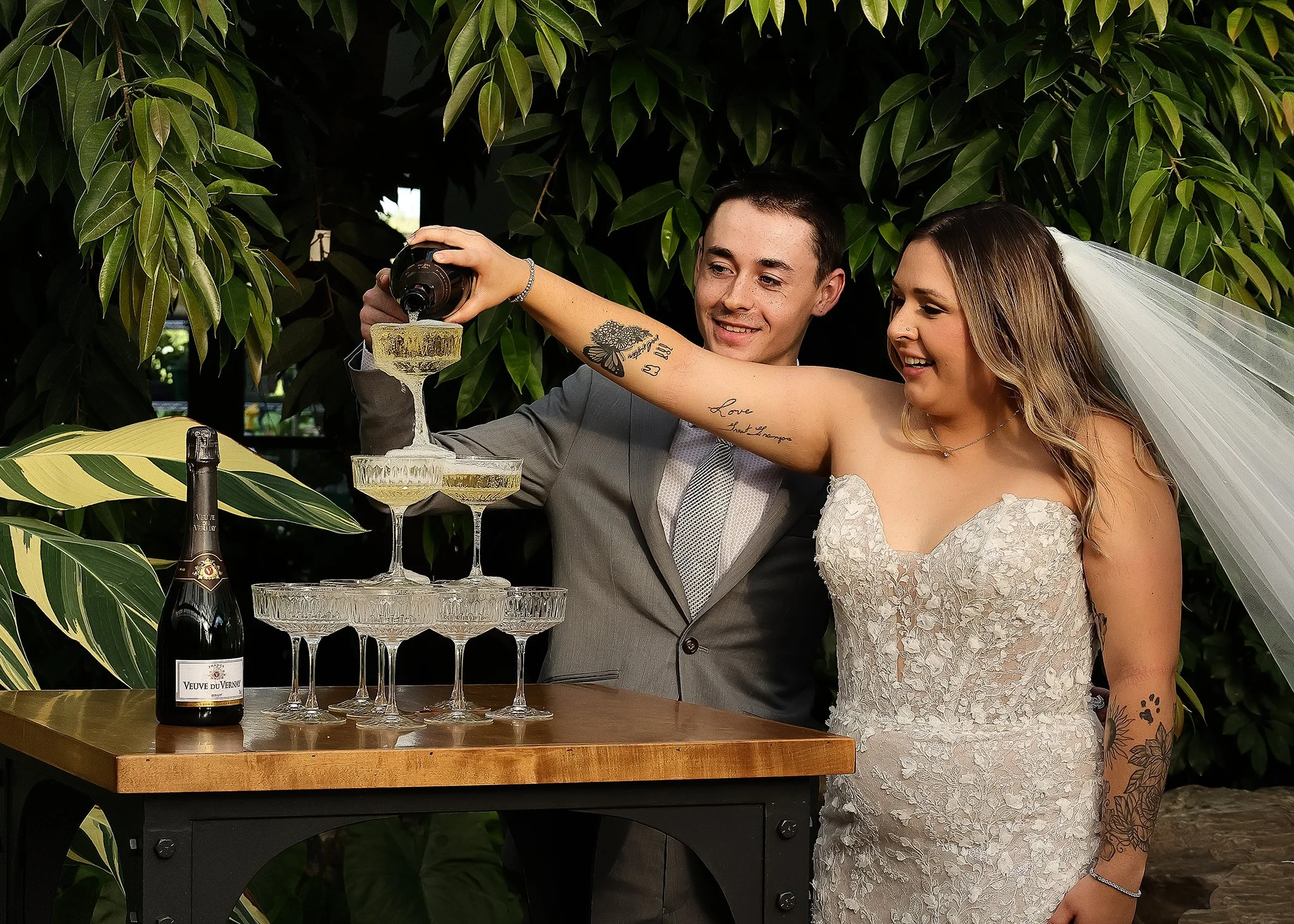 Bride and groom pouring champagne into a tower of glasses outdoors.