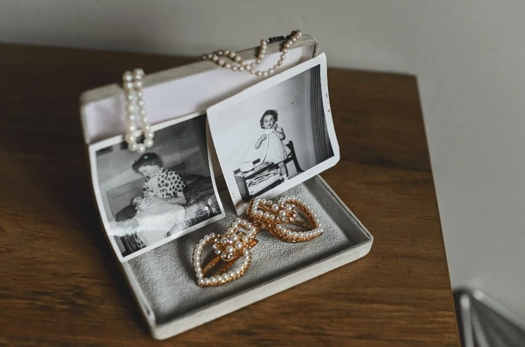 Vintage black and white photographs in a jewelry box with pearl necklace and ornate pearl-embellished hair clips on a wooden surface.