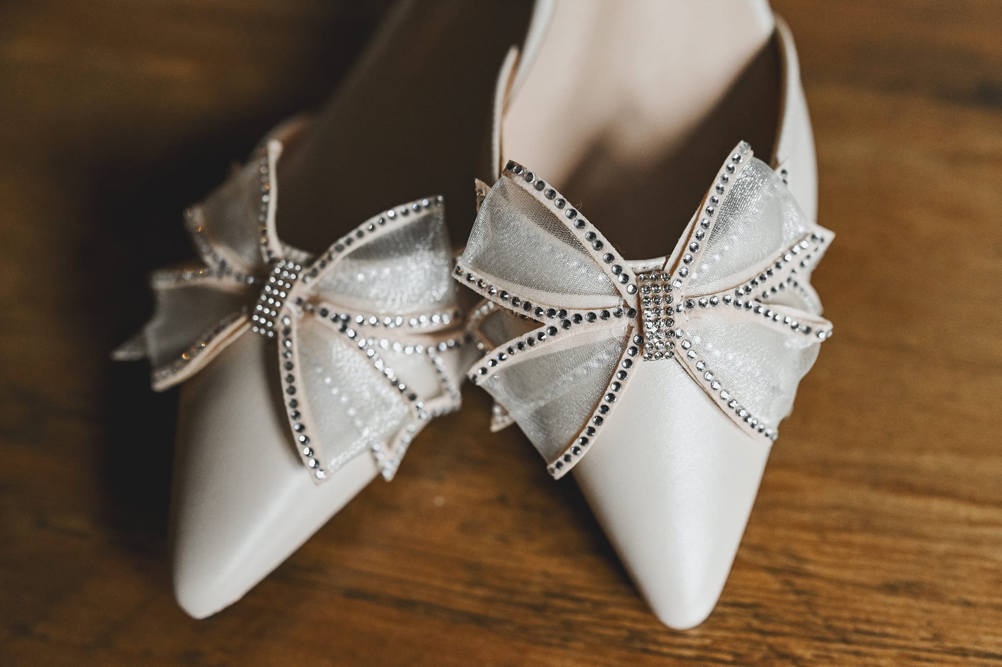 Close-up of elegant white pointed shoes with decorative bow and rhinestones on a wooden surface.