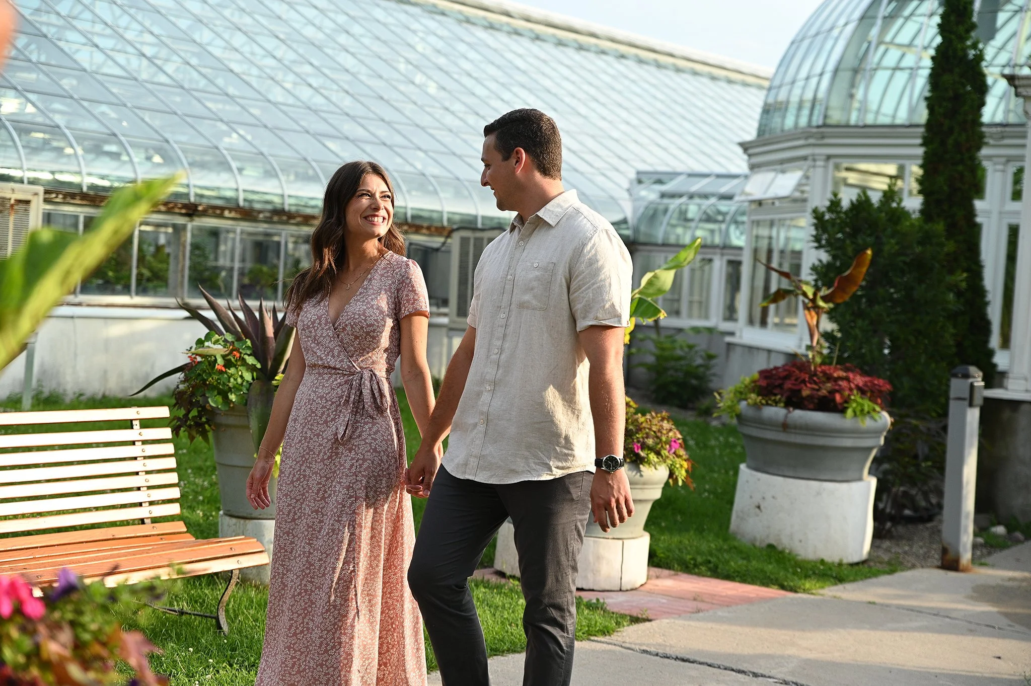 A couple holding hands and smiling at each other while walking in a garden with glass greenhouses in the background.