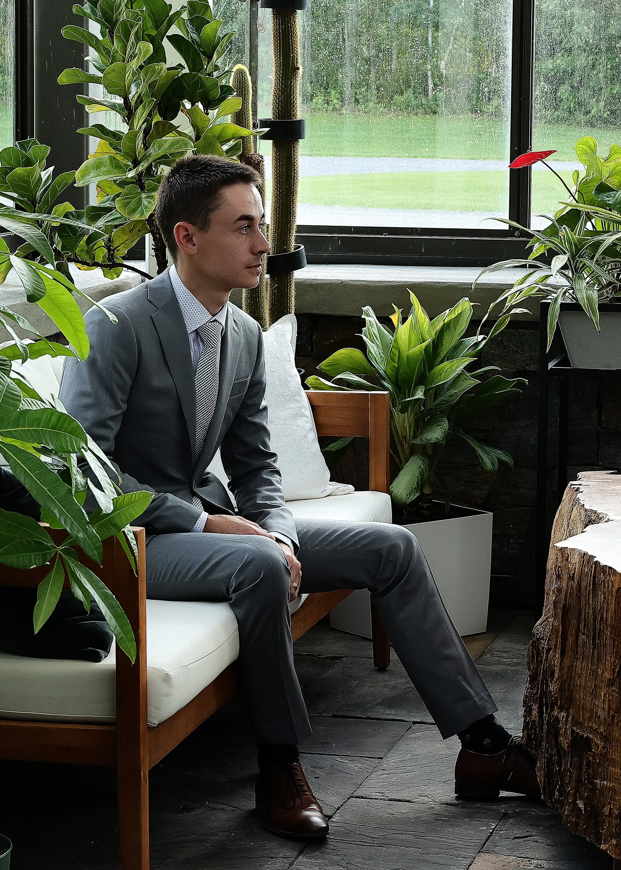Man in gray suit sitting on a bench surrounded by indoor plants in a sunlit room.