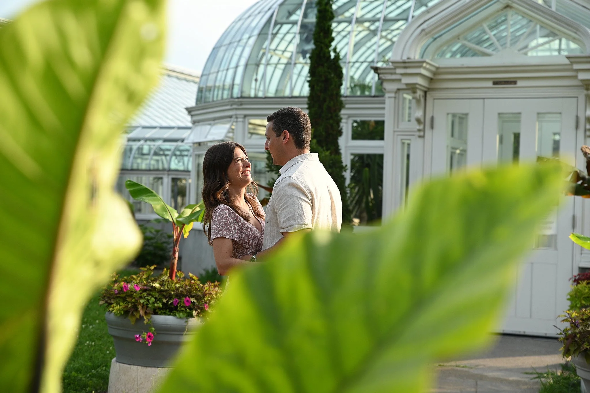 A couple smiling at each other in front of a glass conservatory, surrounded by potted plants and large leaves.
