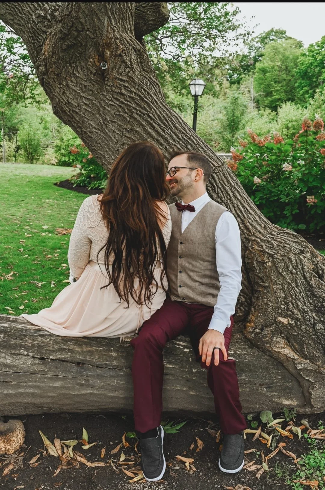 A couple sitting on a tree, smiling at each other, in a park with greenery and a lamp post in the background.