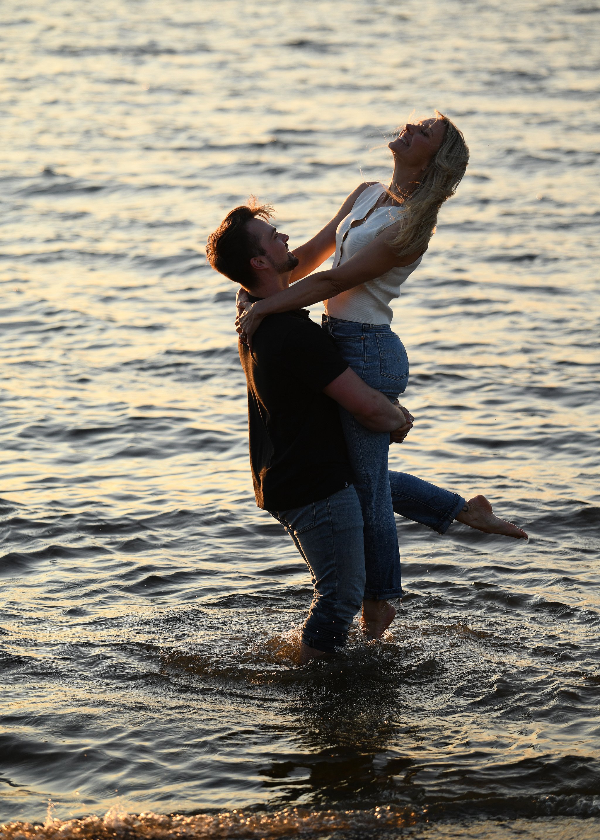 A happy couple playing in the water at sunset. The man is lifting the woman, and both are smiling, enjoying a romantic moment. The water is gently rippling around them.