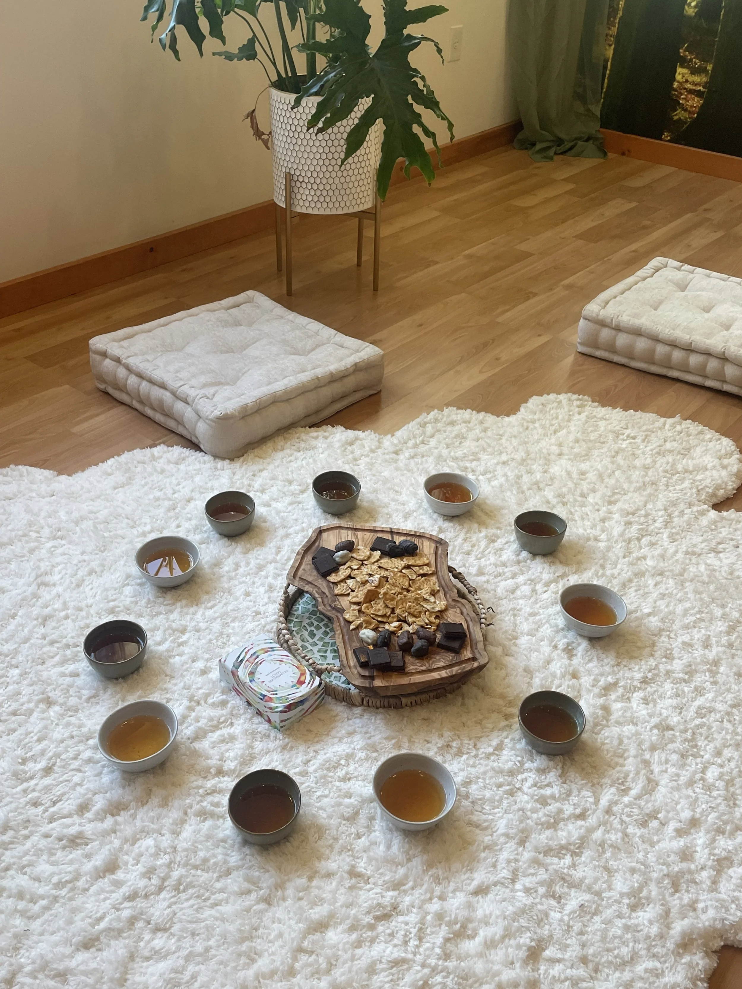 A cozy indoor setting with a large fluffy white rug, around which are nine small cups of tea, a wooden serving tray with cookies and chocolates, and a tissue box. In the background, a potted plant in a decorative white pot and a window with green curtains are visible.