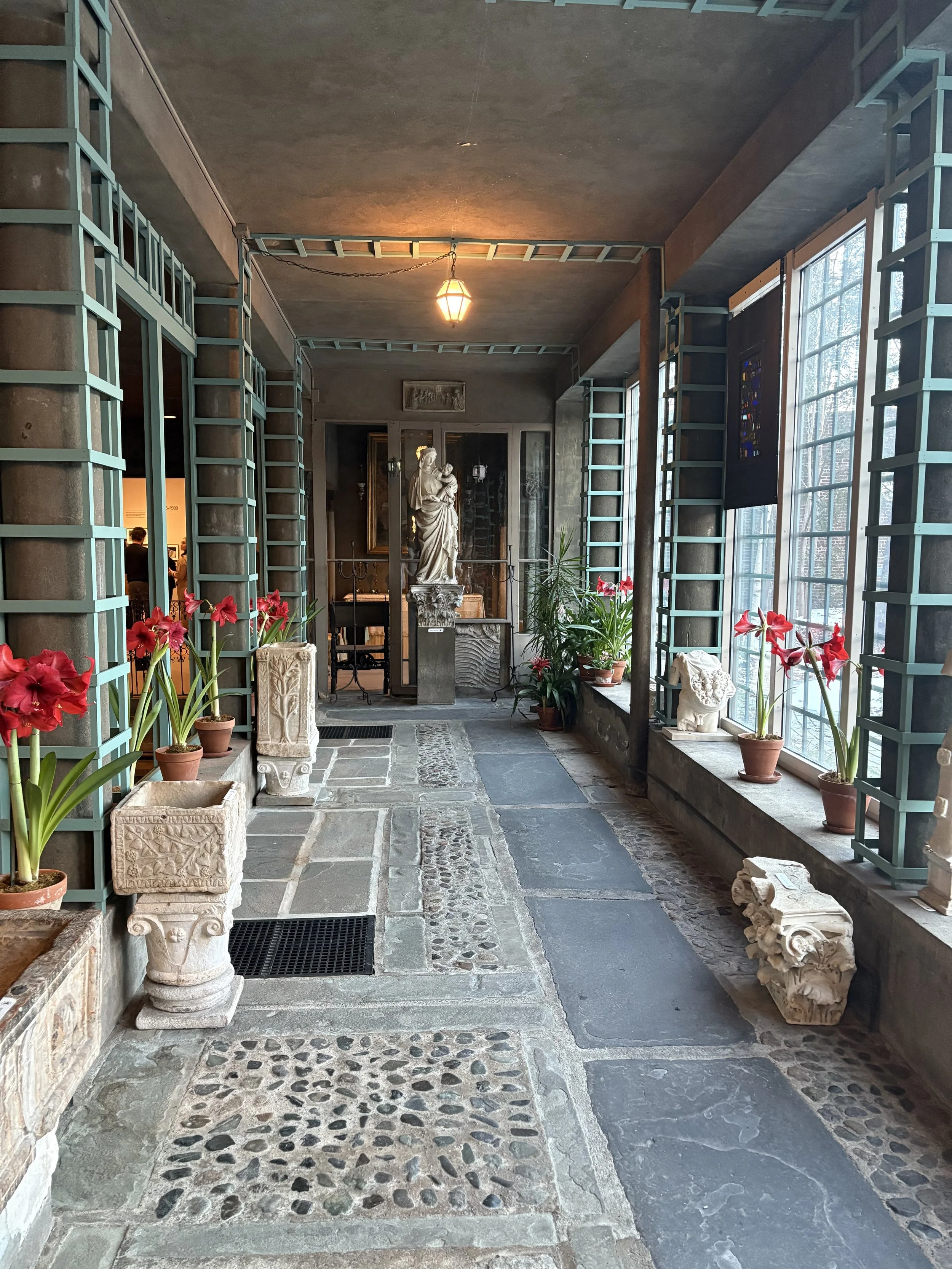 Indoor corridor with large windows on the right, potted plants, classical statues, decorative stone elements, and a rustic stone floor.