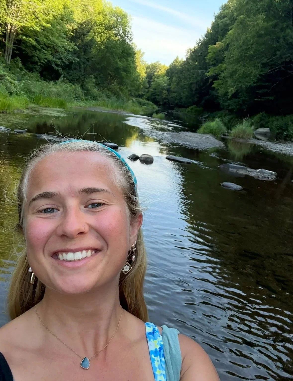 A young woman with blonde hair, wearing earrings, a necklace, and a blue headband, smiling in front of a river surrounded by green trees.
