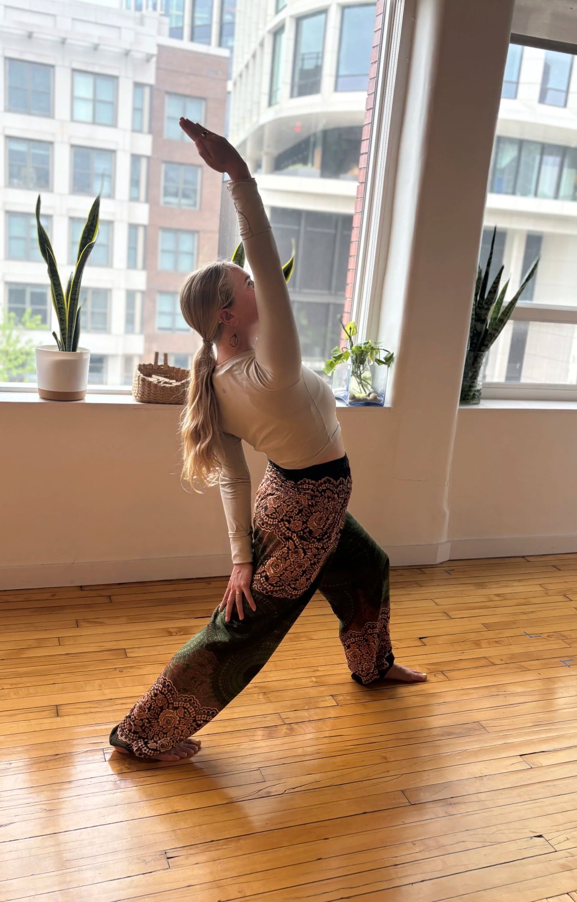 A woman practicing yoga indoors near a large window with city buildings outside, wearing patterned wide-leg pants and a long sleeve top, with her right hand on her thigh and her left arm extended upwards.