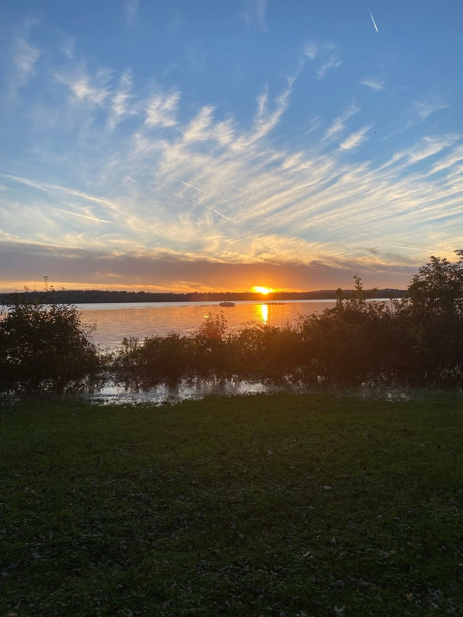 Sunset over a calm river with trees in the foreground and a partly cloudy sky with streaked clouds.