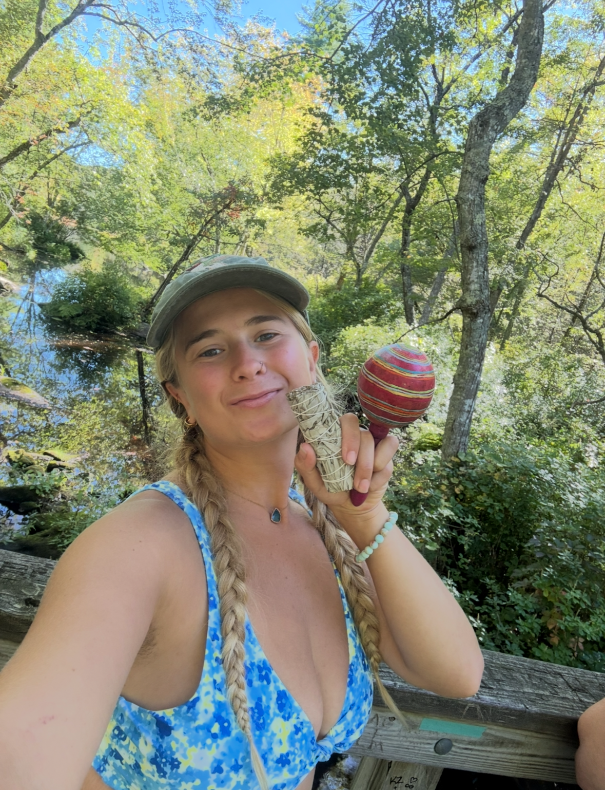 Young woman with blonde hair in braids, smiling while holding a handmade maraca in an outdoor wooded area near a stream.