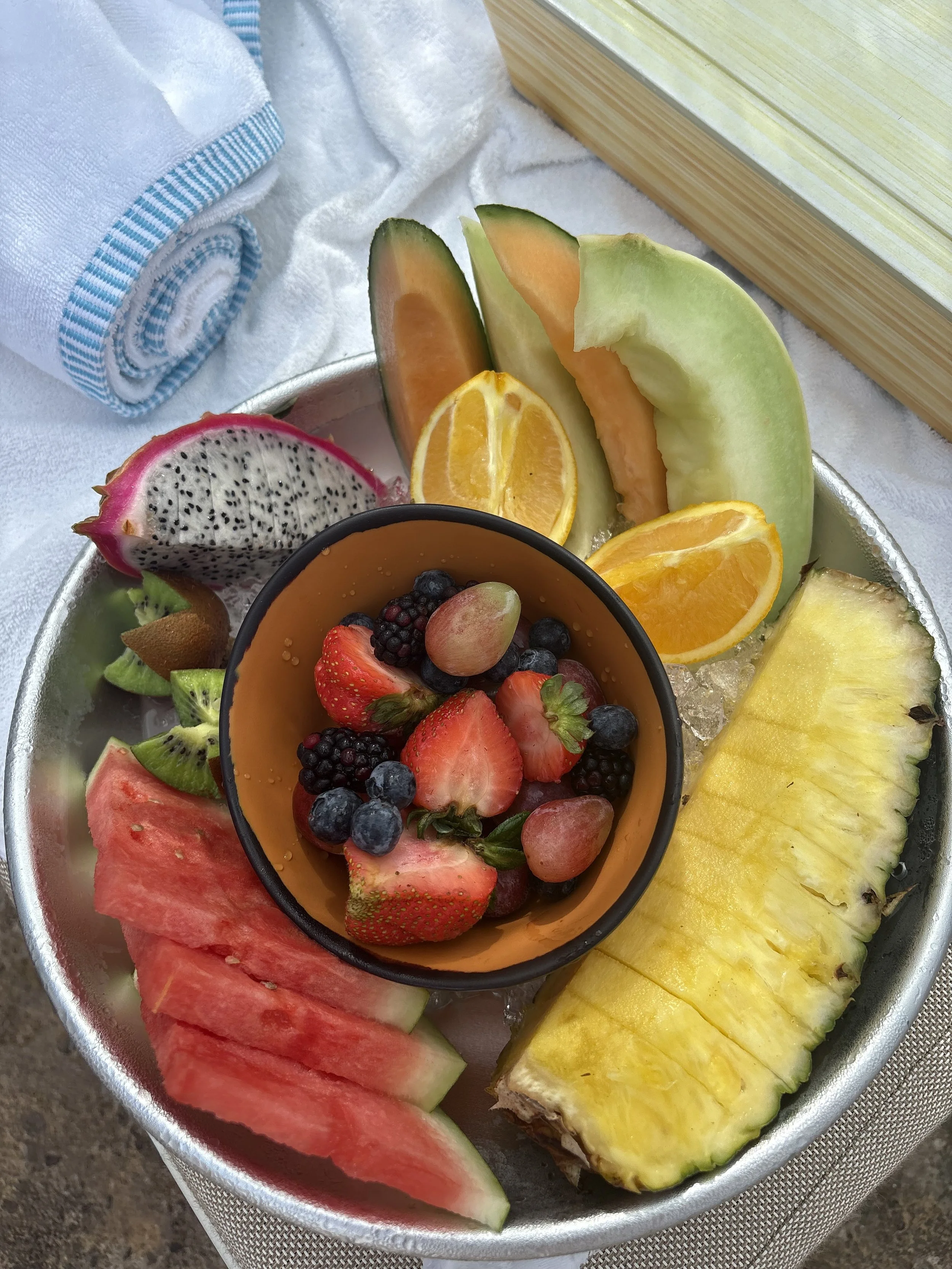 A bowl of sliced fresh fruits including watermelon, kiwi, strawberry, orange, pineapple, cantaloupe, honeydew, dragon fruit, and a small cup of mixed berries, arranged on a bed of ice.
