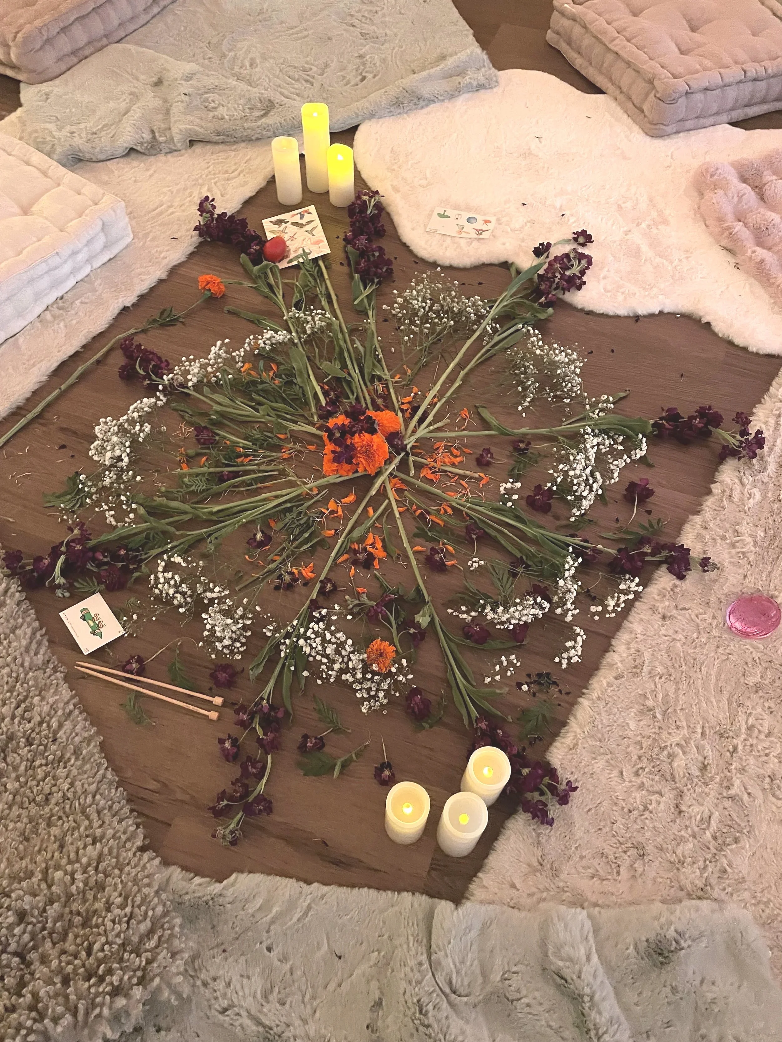 A floral prayer or meditation setup with flowers, candles, and cards on a wooden surface surrounded by various plush rugs and blankets.