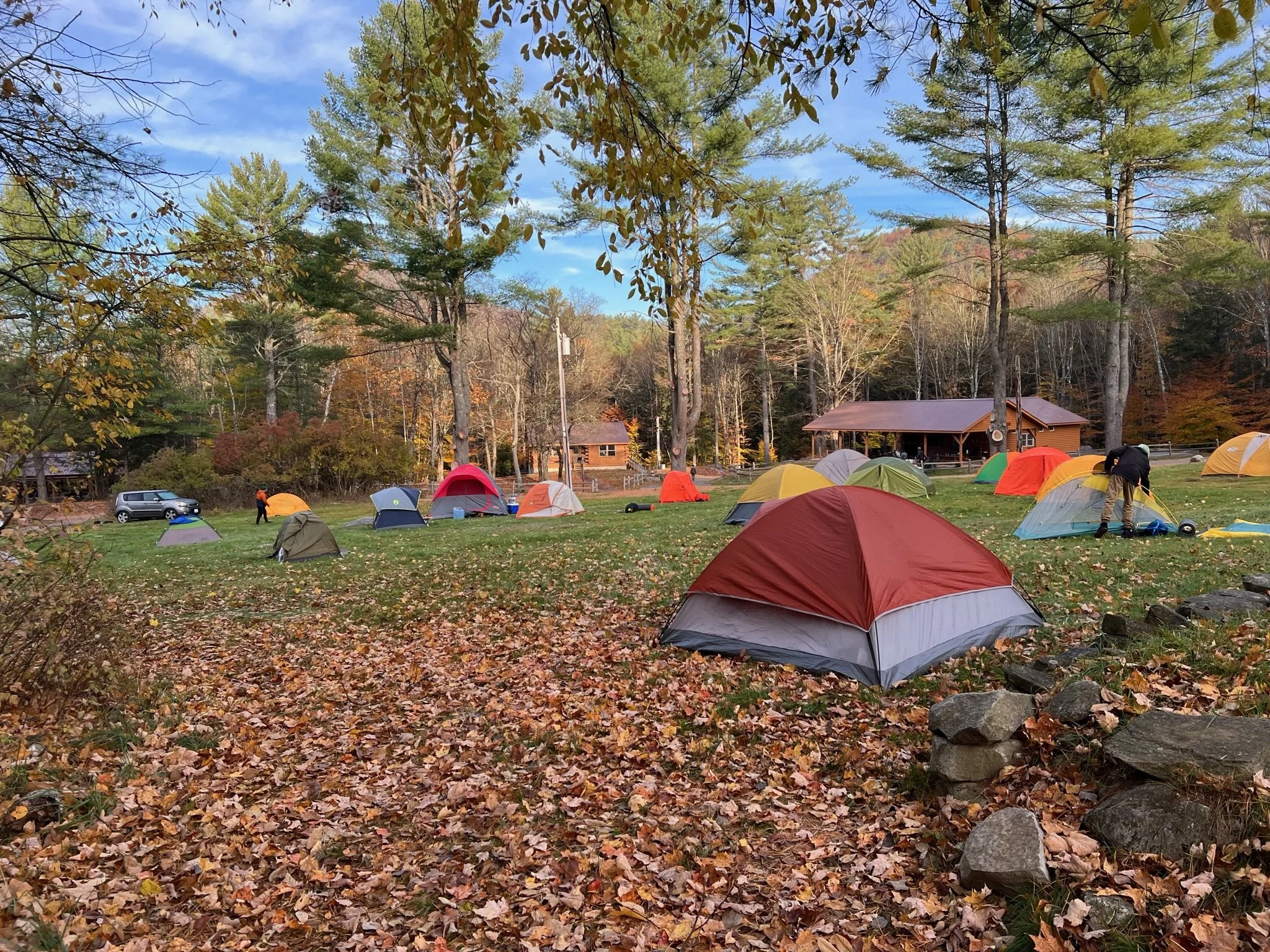 Tent camping area in a forested park during autumn, with several colorful tents set up on a grassy field, fallen leaves on the ground, trees in the background, and a building in the distance.  seasonal wellness cohort, cyclical living support