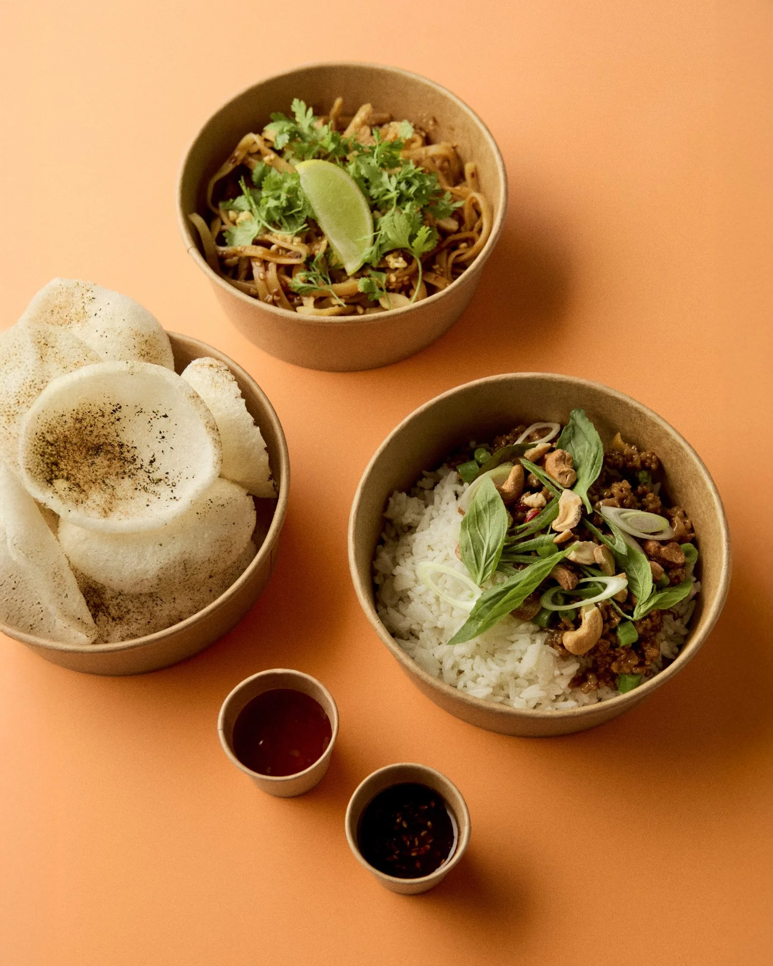 Billede af tre skåle til street food med asiatisk mad, inklusive ris med kød og grøntsager, nudelsalat med lime, og sprøde rispapirruller, med to små skåle med soyasauce og chilisauce.