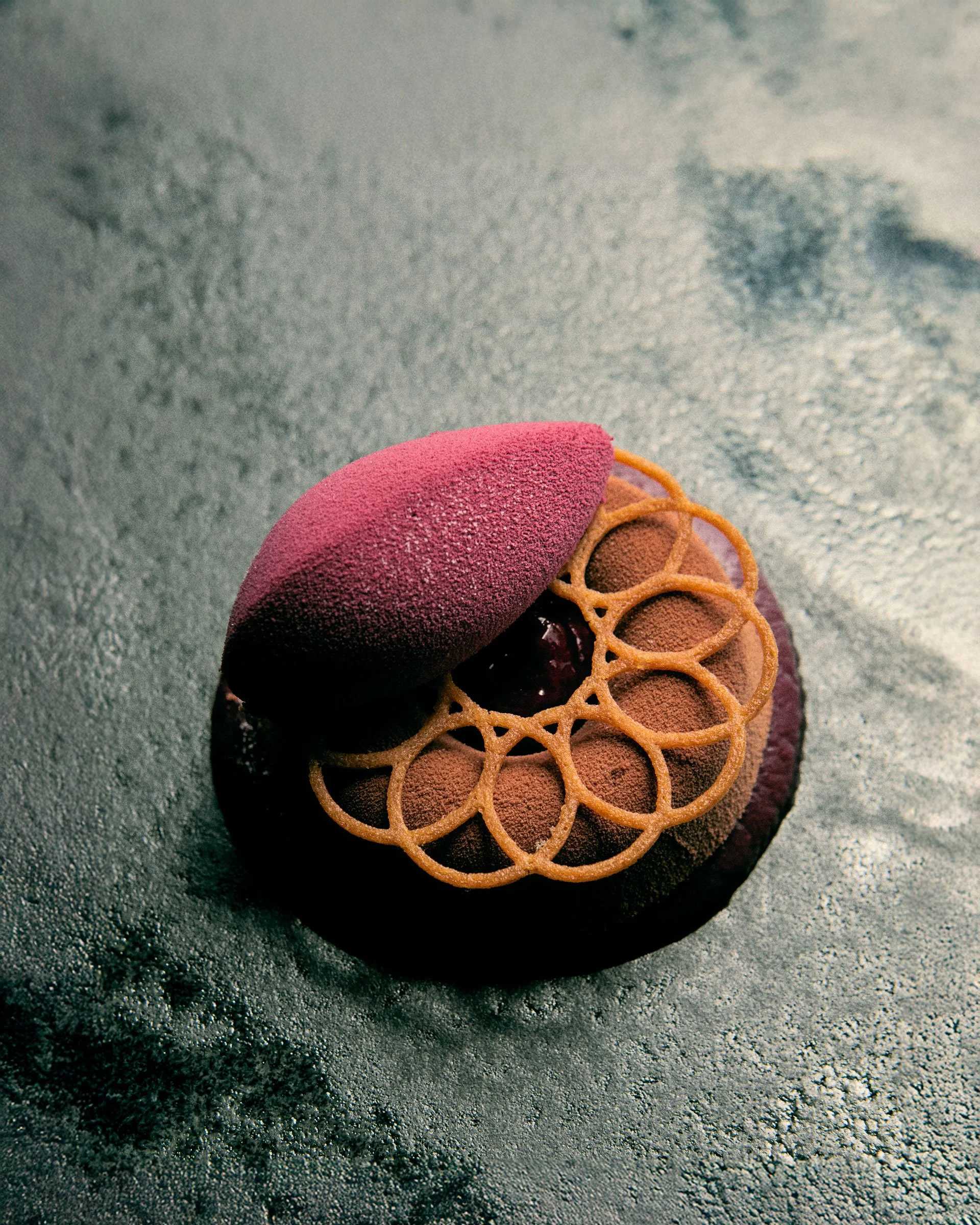 Dekorativ dessert med dyb rød og brun farve, toppet med en pink skum og en fin luftsprængt kage.