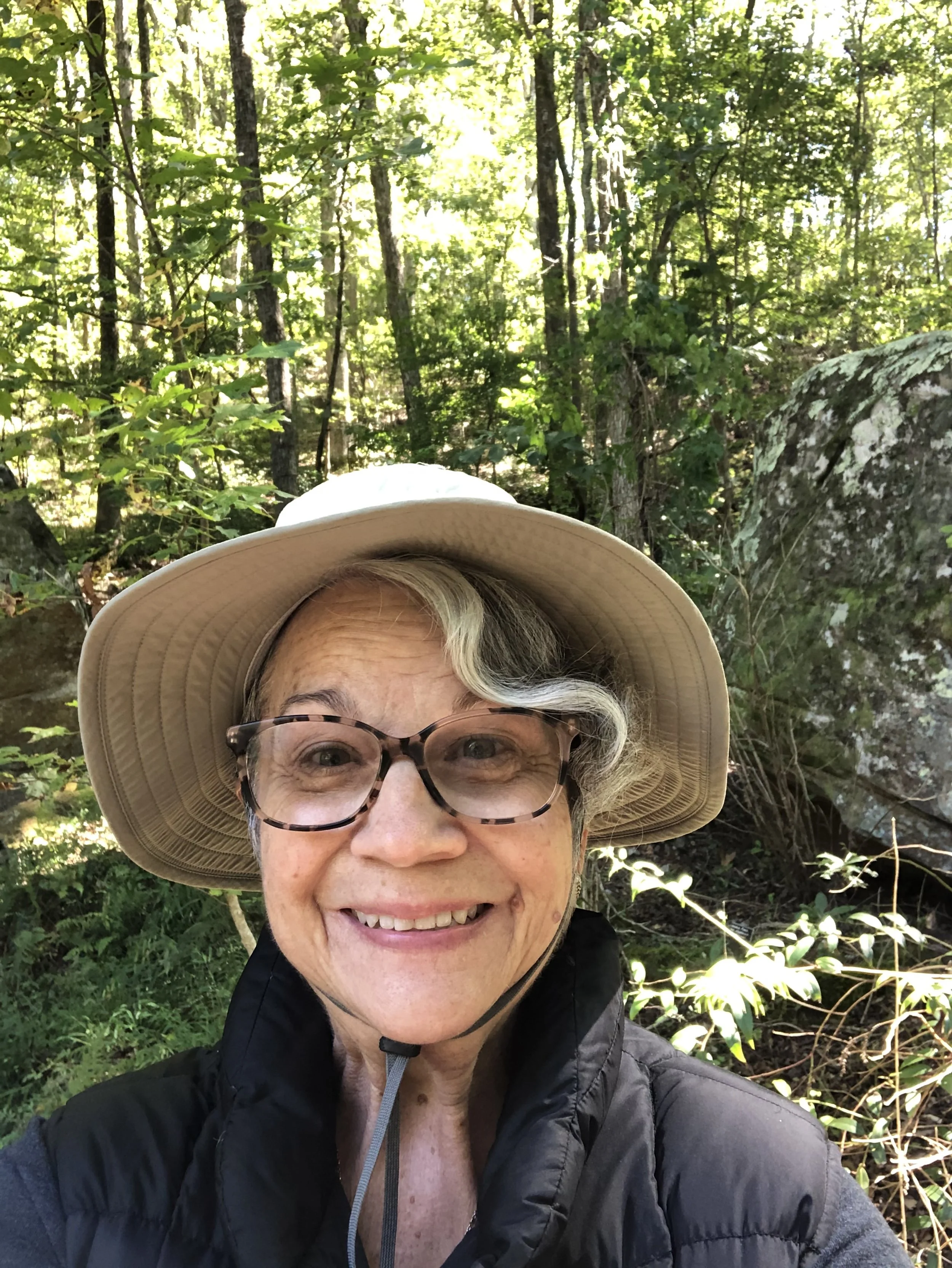 A smiling woman in a wide-brimmed hat and glasses taking a selfie in a green forest.