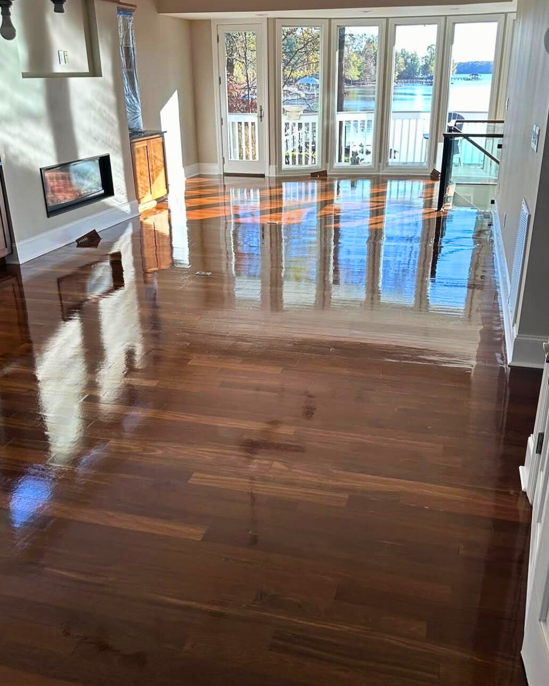 Living room with polished hardwood floors, large windows with a view of a lake and trees, and a balcony.